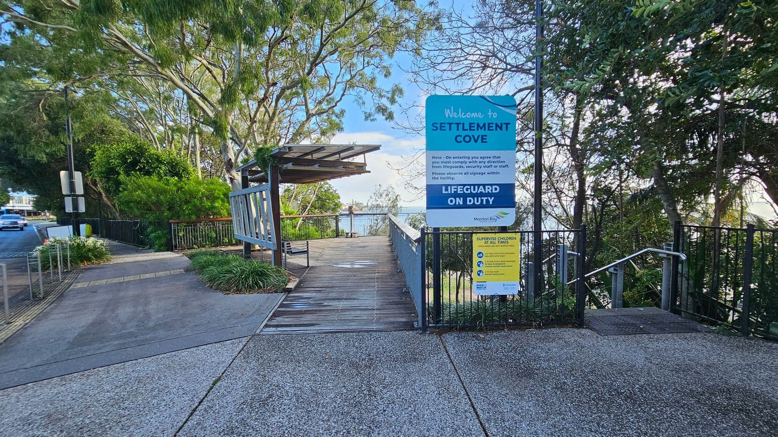 Entrance to Settlement Cove with a large sign on the right detailing rules and lifeguard information. A pathway leads downwards, flanked by greenery and trees. On the left, a road with a parked car and more trees. A wooden structure with a bench is visible near the centre. The background shows glimpses of water through the trees. The ground is paved with concrete, and there are metal railings along the path.