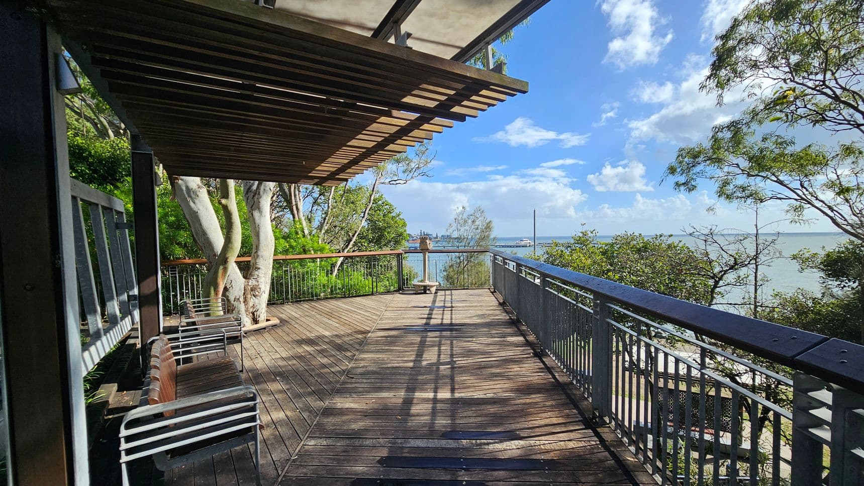 A wooden deck extends into the distance, bordered by a metal railing on the right. On the left, there are several metal and wood chairs under a slatted wooden awning. Trees with light-coloured trunks and green foliage are visible on the left, while more trees and shrubs are on the right. In the background, a body of water stretches out, with a distant shoreline and a ship visible. The deck is sunlit, casting shadows on the wooden floor.