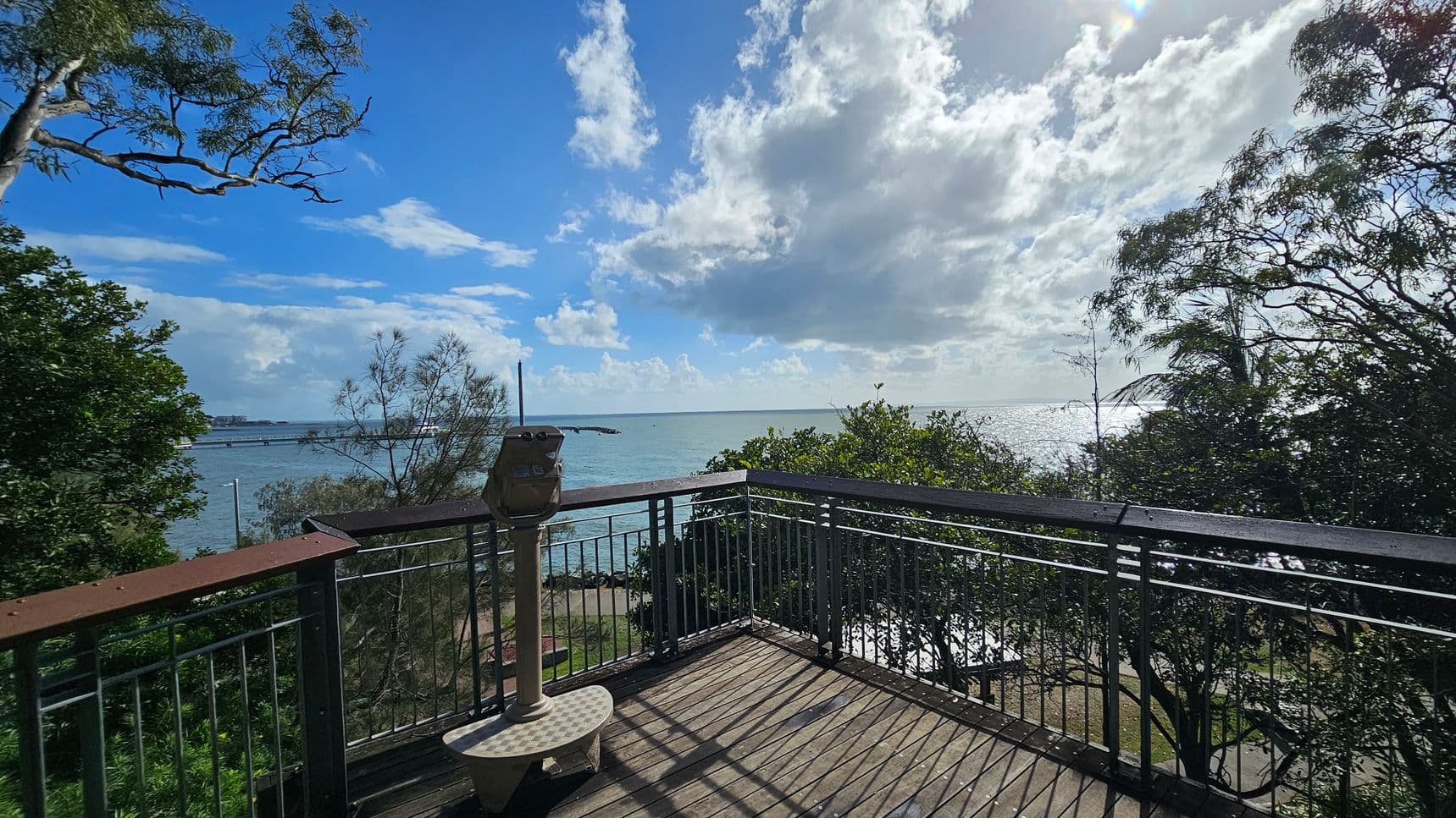 A wooden viewing platform with metal railings overlooks a body of water. In the centre, a mounted binocular viewer is positioned on the platform. Trees with sparse foliage frame the scene on the left and right. The water stretches out to the horizon, reflecting sunlight. A few clouds are visible in the sky, and a distant shoreline is faintly visible on the left. The platform floor is wooden, casting shadows from the railings.