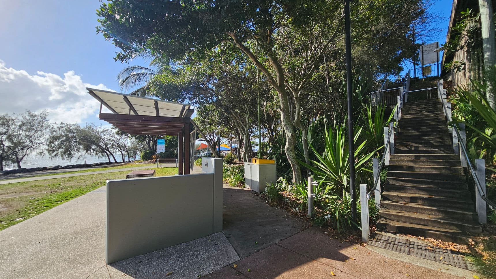 A pathway leads to a covered seating area on the left, with a bench and a sign nearby. Trees and shrubs line the path, providing shade. On the right, a set of stairs ascends, bordered by lush green plants and a railing. The background features a glimpse of the sea and more trees, while the foreground shows a paved walkway. A yellow-topped bin is visible near the stairs.