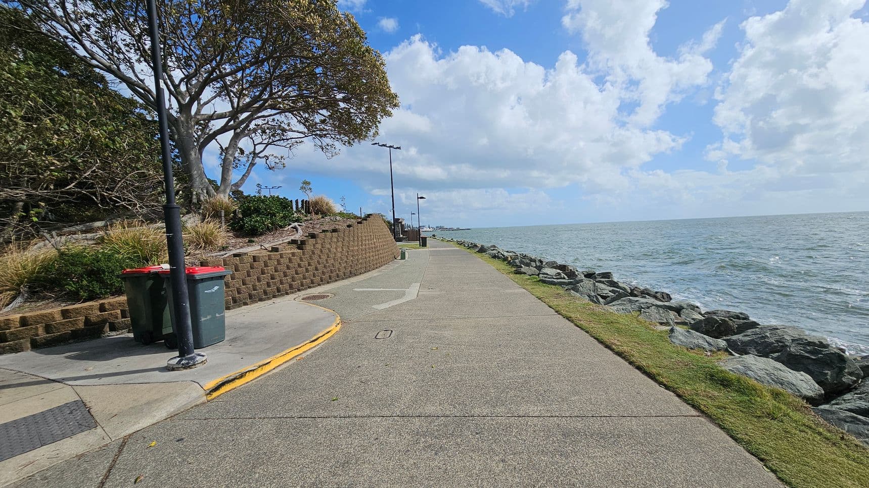 A coastal pathway runs alongside the ocean on the right, bordered by large rocks. On the left, a brick retaining wall supports a grassy area with a large tree and shrubs. A lamppost stands near two rubbish bins on a concrete section of the path. The path is wide and paved, with a slight curve. The ocean is calm, and the sky is filled with scattered clouds.