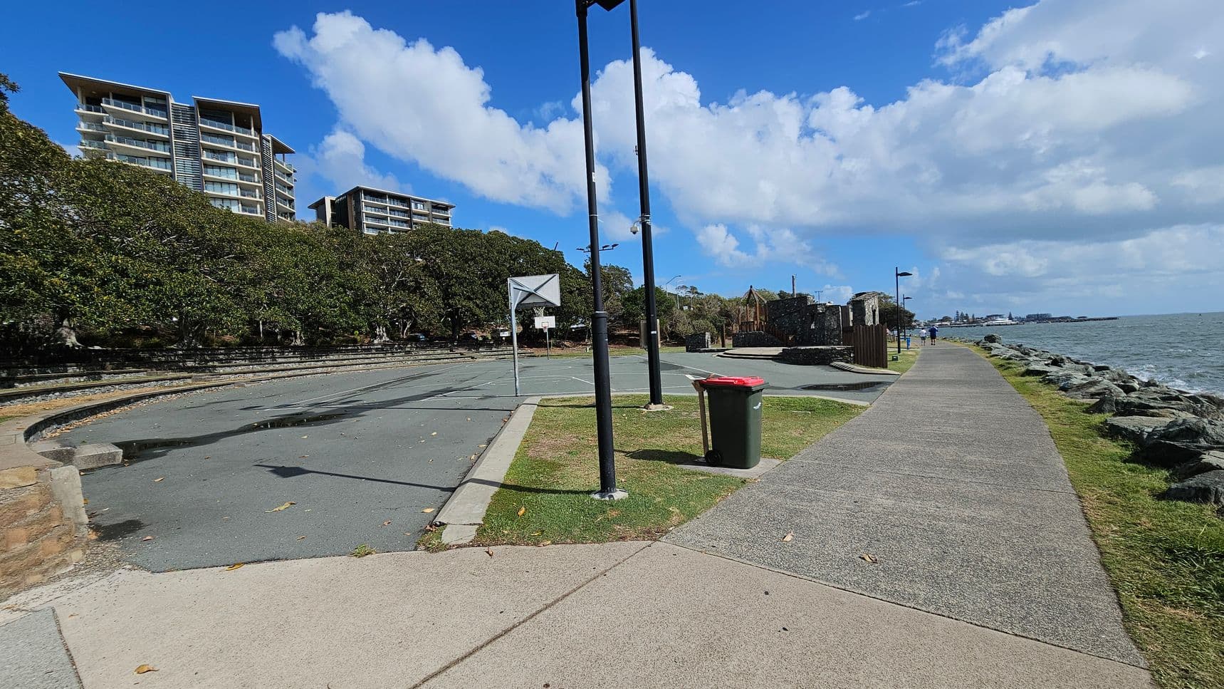 A coastal pathway runs along the right side, bordered by rocks and the sea. On the left, there are tall residential buildings behind a row of dense trees. A basketball court with a hoop is visible, and a red-lidded bin stands near a tall lamp post. The ground is a mix of concrete and grass, and the sky is partly cloudy.