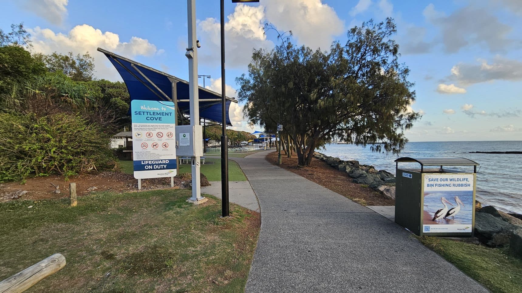 A coastal pathway with a sign on the left reading "Welcome to Settlement Cove" and "Lifeguard on Duty." Nearby, a blue-roofed shelter and picnic tables are visible. On the right, a bin with a sign urging to "Save Our Wildlife, Bin Fishing Rubbish" is placed near the rocky shoreline. Trees line the path, which curves along the water's edge. The sky is partly cloudy.