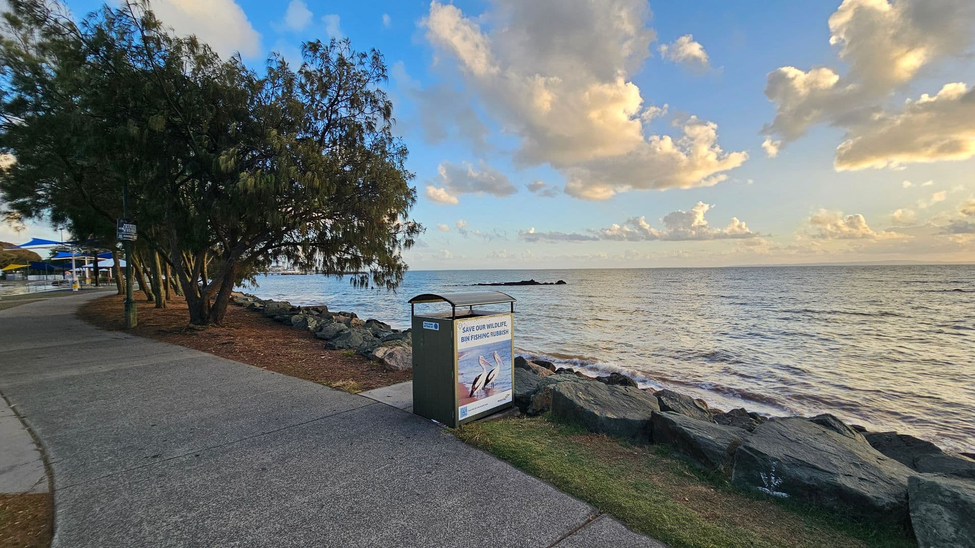 A coastal pathway runs along the left side, bordered by trees and a grassy area. A bin with a sign about wildlife conservation is positioned near the path. Large rocks form a barrier between the path and the sea on the right. The ocean extends to the horizon, with gentle waves visible. The sky is filled with scattered clouds, reflecting soft light on the water.