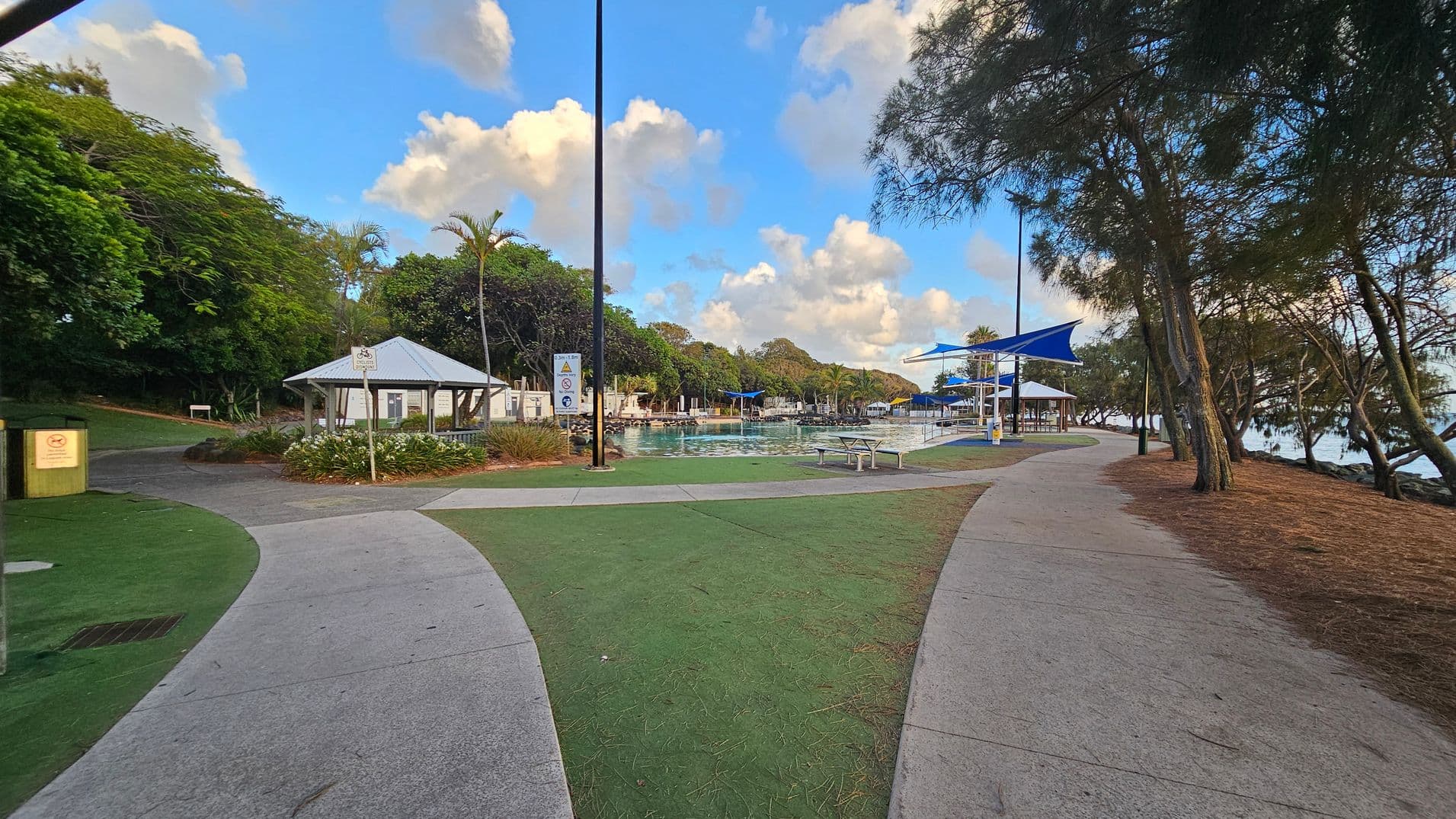 A park with a paved path splitting into two directions. On the left, there's a small building with a white roof surrounded by lush green trees and a bin nearby. In the centre, a pool with blue shade structures and benches is visible. On the right, trees line the path, leading towards a body of water. The ground is a mix of concrete and green grass, with some pine needles scattered around.