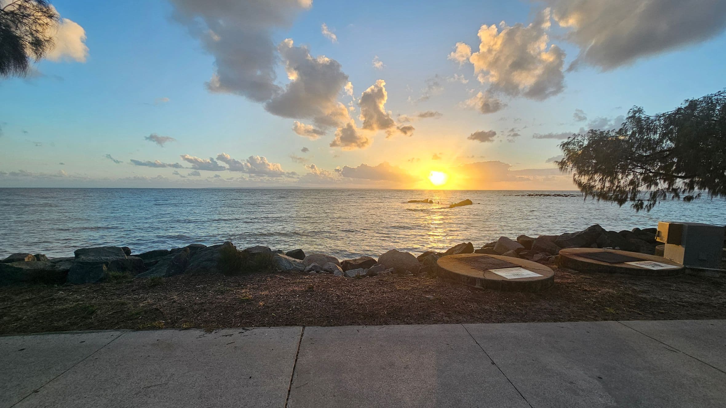 A coastal scene with a concrete path in the foreground leading to a rocky shoreline. The sun is setting on the horizon, casting a warm glow over the ocean. On the right, there are two circular structures and a small bin. Trees with sparse foliage frame the image on the left and right. The sky is partly cloudy, with the clouds reflecting the sunlight.