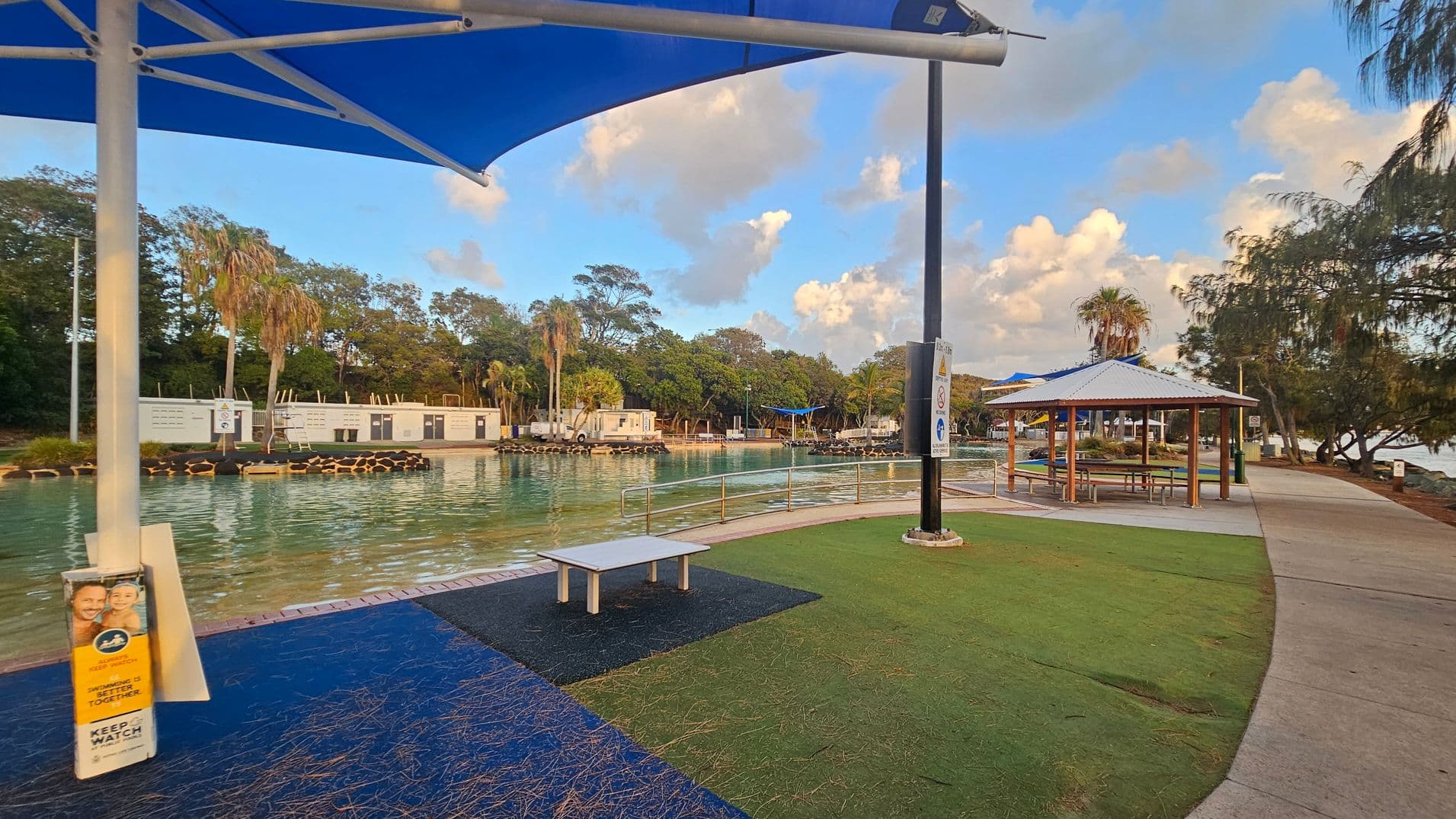 A waterfront area with a paved path on the right, bordered by green grass. A wooden gazebo with a metal roof and picnic tables is near the path. A blue shade structure is visible on the left, above a bench. The water is calm, with a few small boats docked near a building in the background. Trees line the far side of the water, and a sign is attached to a pole in the foreground.