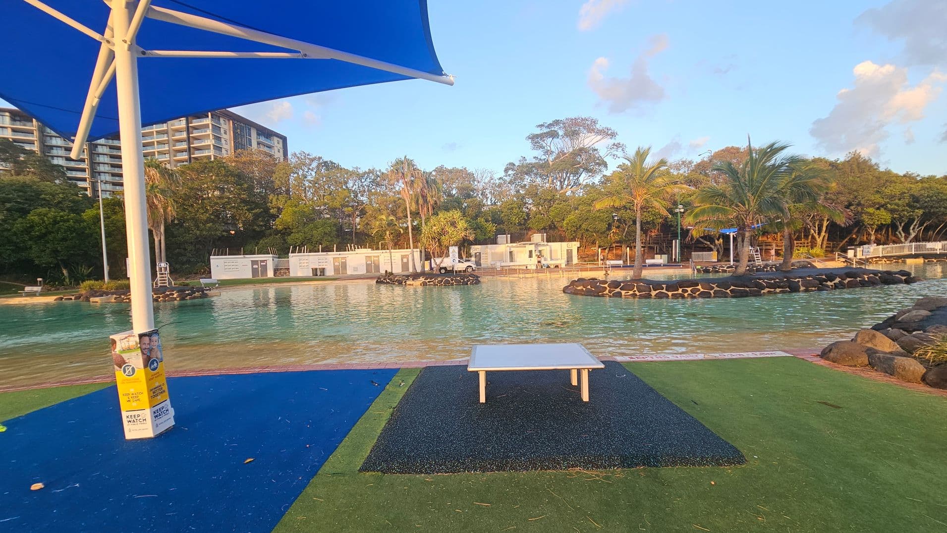 A pool area with a blue sunshade on the left, covering a section of blue and green flooring. A white table is in the centre on a black mat. The pool is bordered by rocks and surrounded by palm trees and greenery. In the background, there are white buildings and a tall apartment complex. The scene is lit by warm sunlight.