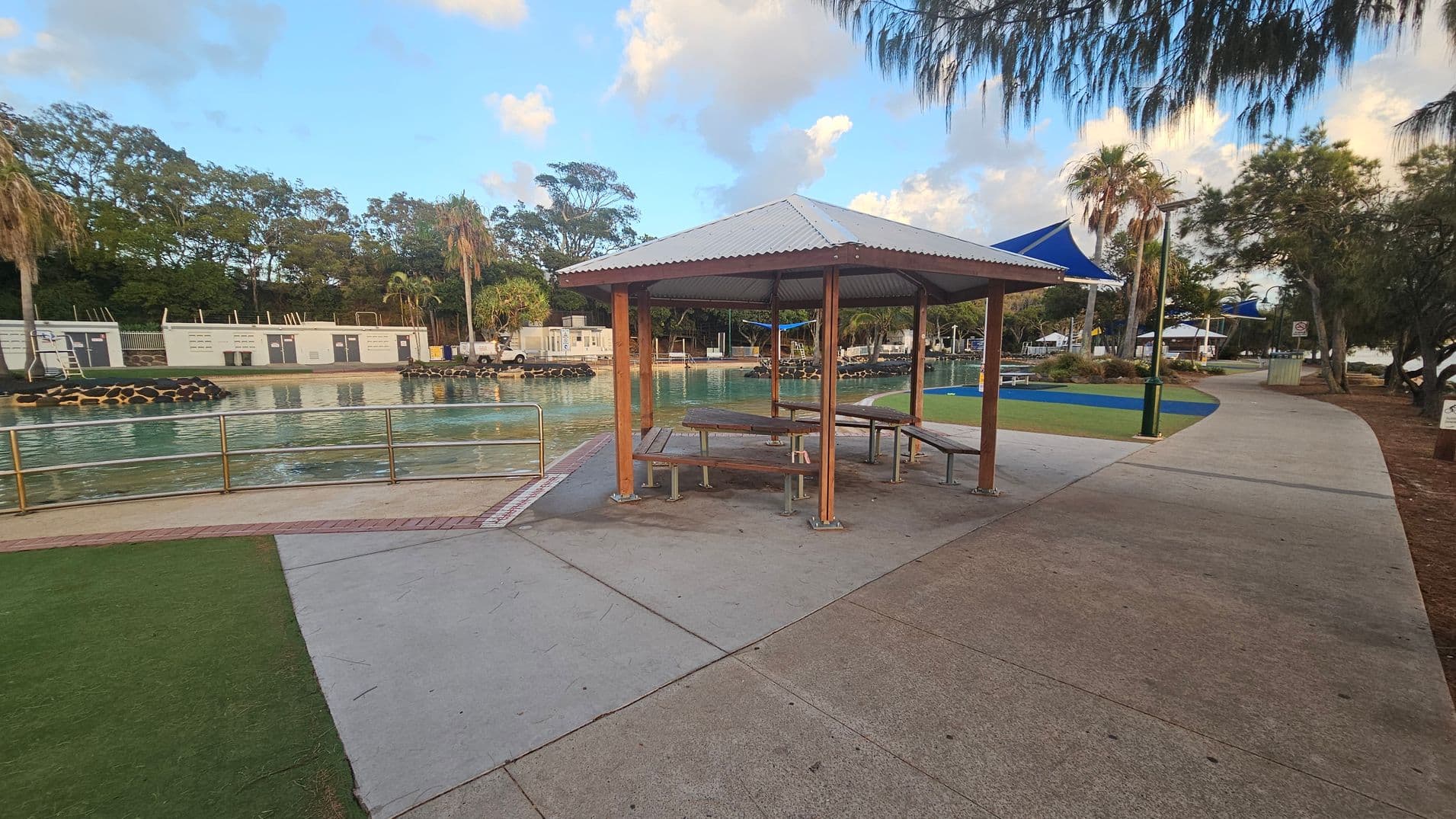 A park area with a covered picnic shelter in the centre, featuring wooden benches and a table. To the left, there's a fenced water area with a railing and some buildings in the background. The right side has a paved pathway curving through the park, lined with trees and additional seating areas under blue shade structures. The ground is a mix of concrete and grass, and there are palm trees scattered throughout the scene.
