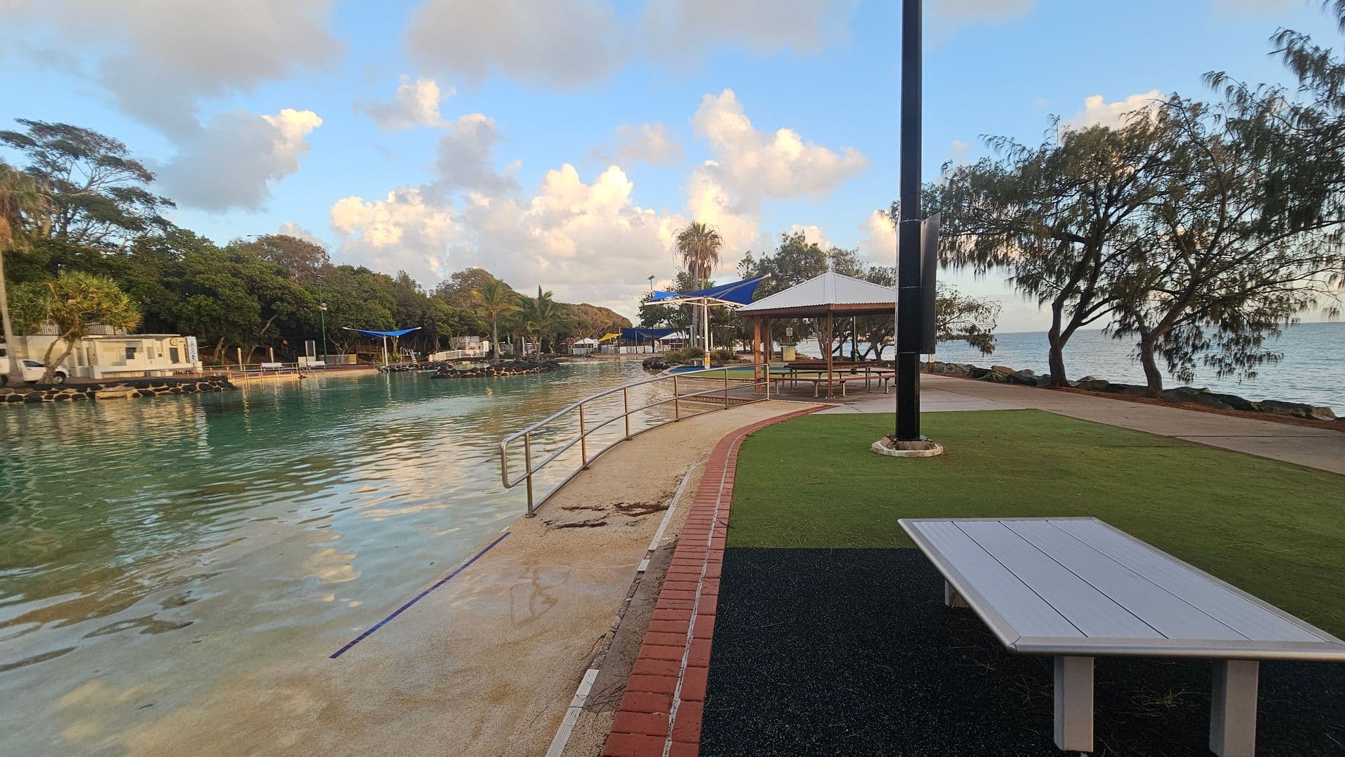 A waterfront area with a curved pathway bordered by a red brick line. On the right, there's a grassy area with a white picnic table and a large black pole. A gazebo with a white roof and benches is nearby. Trees line the water's edge, and the water is calm with a slight reflection of the sky. In the background, more trees and a few structures are visible, including a blue shade structure. The scene is peaceful and open.