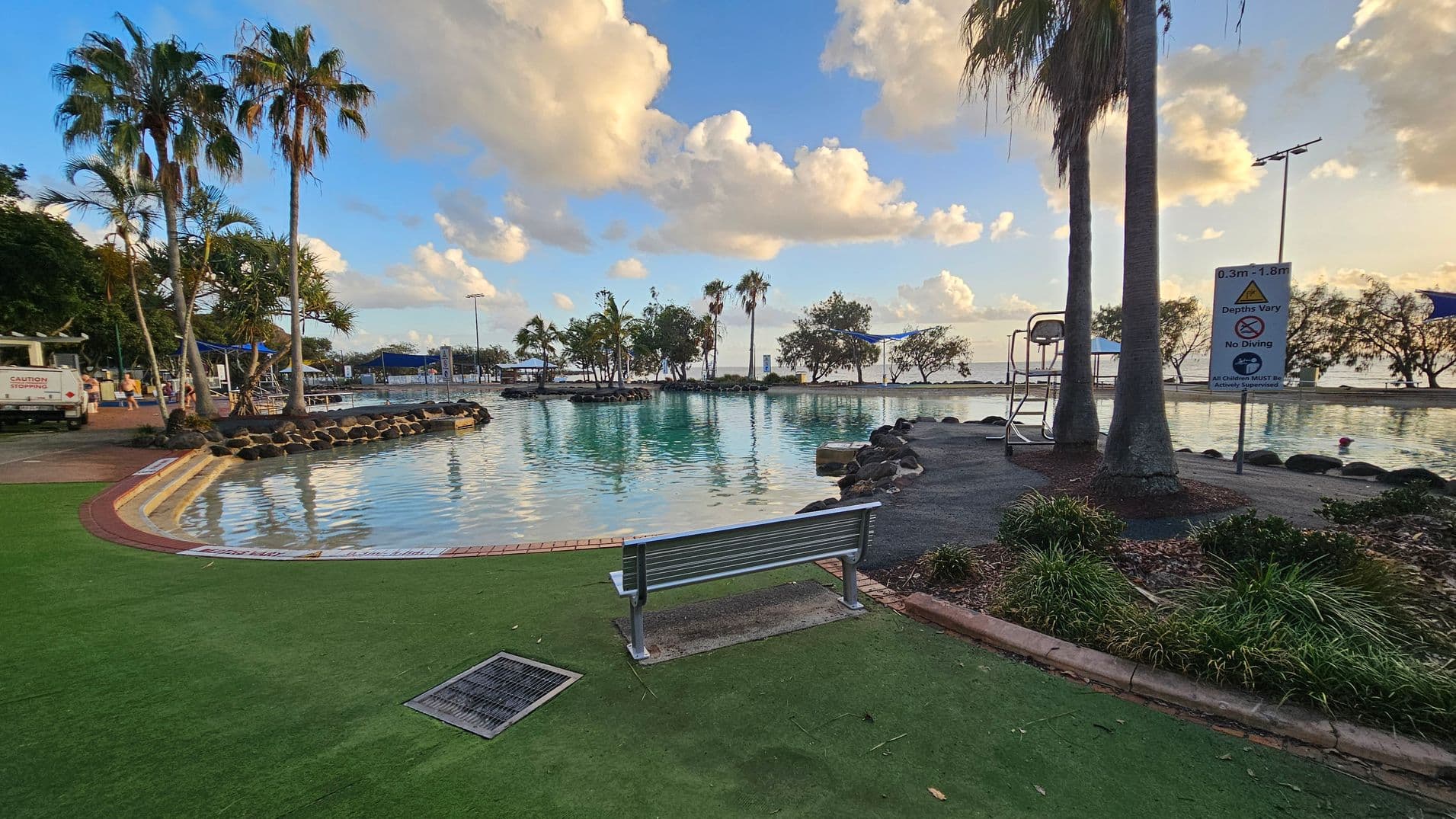 A tranquil pool area with a bench in the foreground on a green artificial turf surface. To the right, a sign indicates varying water depths and no diving. Palm trees and other tropical plants are scattered around, with a lifeguard chair nearby. The pool is bordered by rocks, and in the background, more palm trees and a glimpse of the sea are visible. The sky is partly cloudy, casting reflections on the water.
