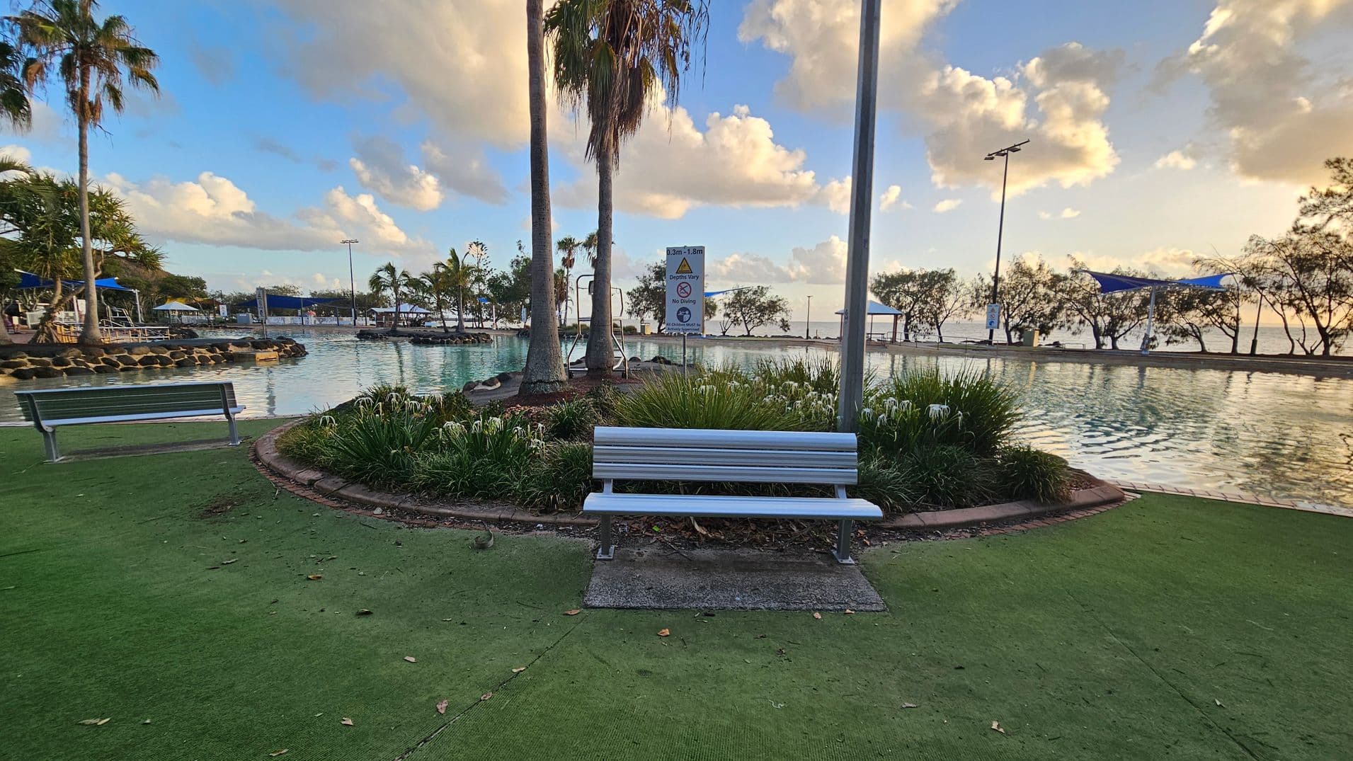 A park scene with a white bench in the foreground, surrounded by green grass and plants. Behind the bench, there is a small body of water with a few palm trees and a sign. On the left, another bench is visible near a rocky edge. In the background, there are more trees and a blue shade structure. The sky is partly cloudy, and the water reflects the light.