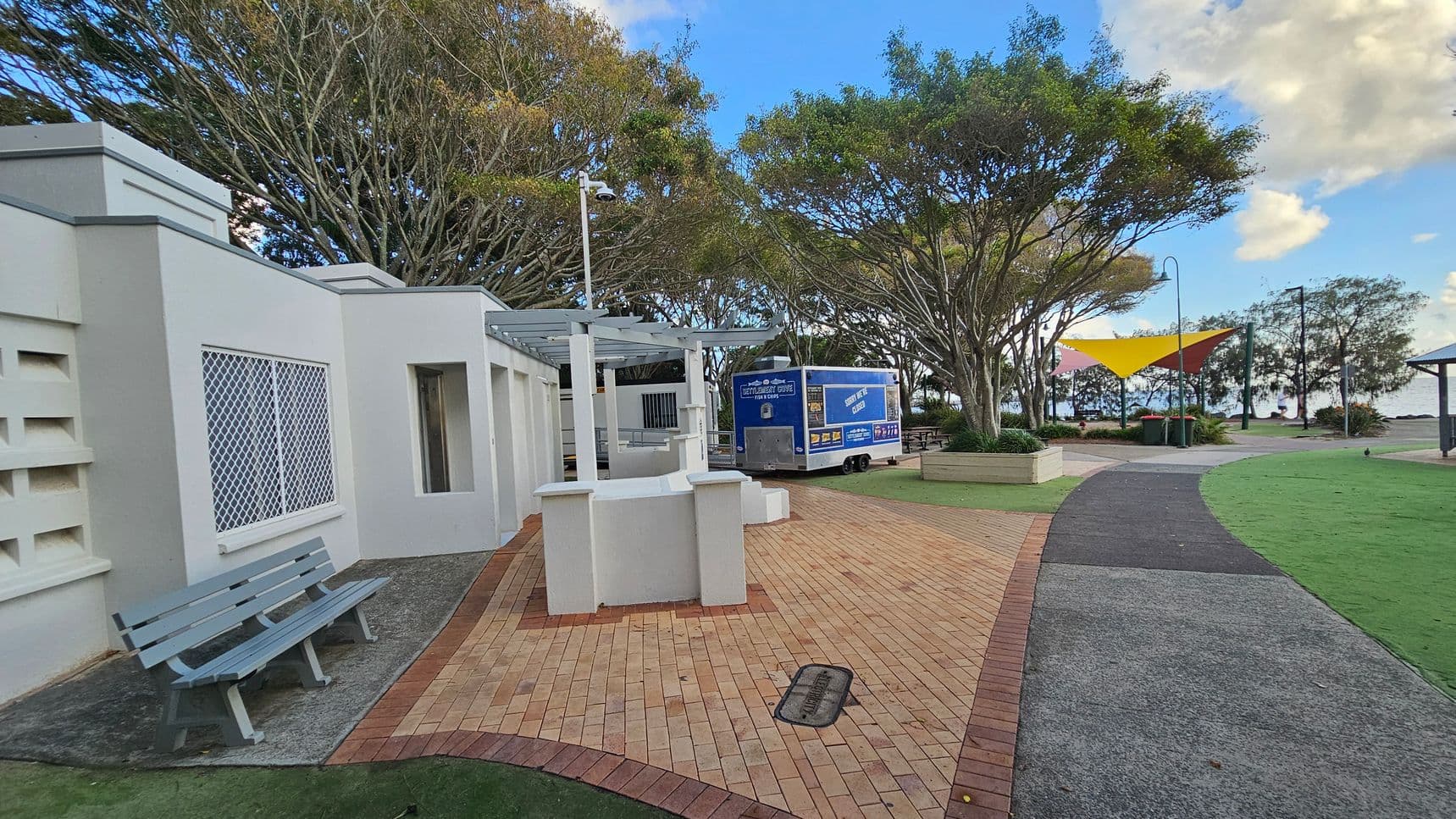 A paved walkway curves through a park area. On the left, there's a light grey building with a blue bench in front. A blue food truck is parked nearby. Trees with dense foliage line the background, providing shade. On the right, a yellow and red triangular shade structure is visible above a seating area. The ground is a mix of brick paving and green grass, with a path leading towards the water in the distance.