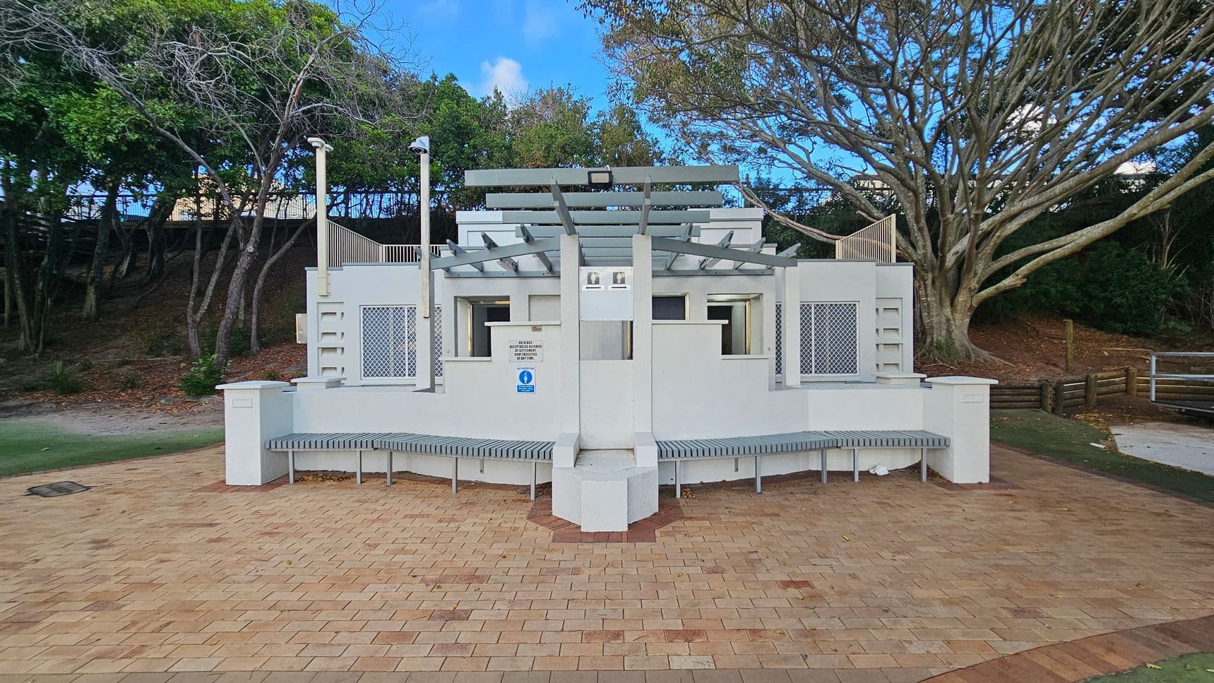 A public restroom facility with a modern design, featuring a white exterior and a central entrance. The structure has a pergola-style roof and is flanked by two benches. The ground is paved with light brown and beige bricks. In the background, there are trees with dense foliage on the left and a large tree with sprawling branches on the right. A small grassy area is visible to the left, and a wooden fence is on the right.