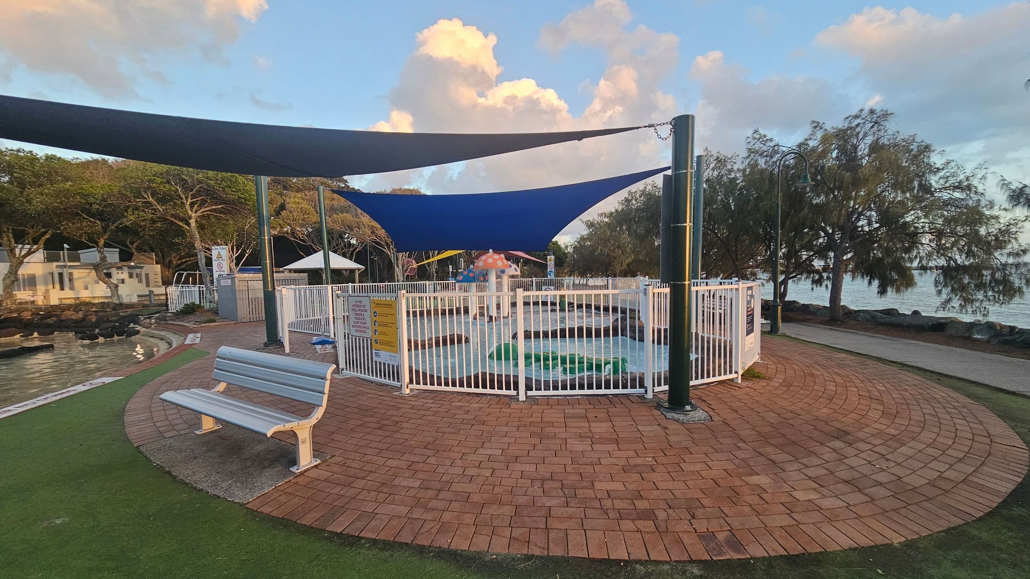 A fenced children's splash pool with a mushroom-shaped water feature is in the centre, covered by blue shade sails. A white bench is on the left, set on a circular brick-paved area with grass surrounding it. Trees line the background, and a pathway runs alongside a body of water on the right.