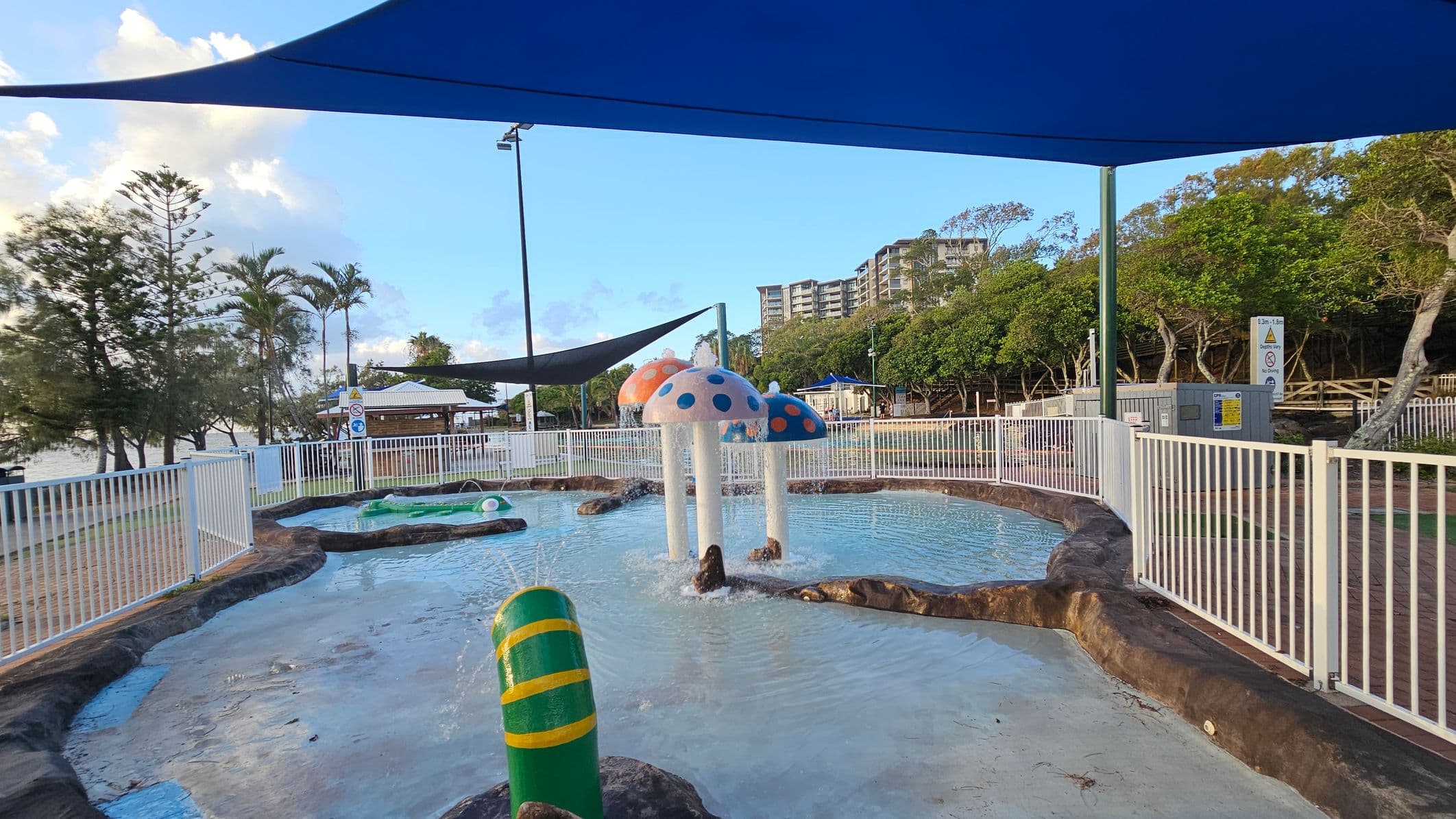 A children's water play area with a shallow pool featuring colourful mushroom-shaped fountains in the centre. The pool is surrounded by a white fence. On the left, there are palm trees and a view of the water. On the right, there are more trees and a sign. A large blue shade cloth covers the area. In the background, there are buildings and a wooden walkway. The ground is a mix of concrete and natural rock formations.