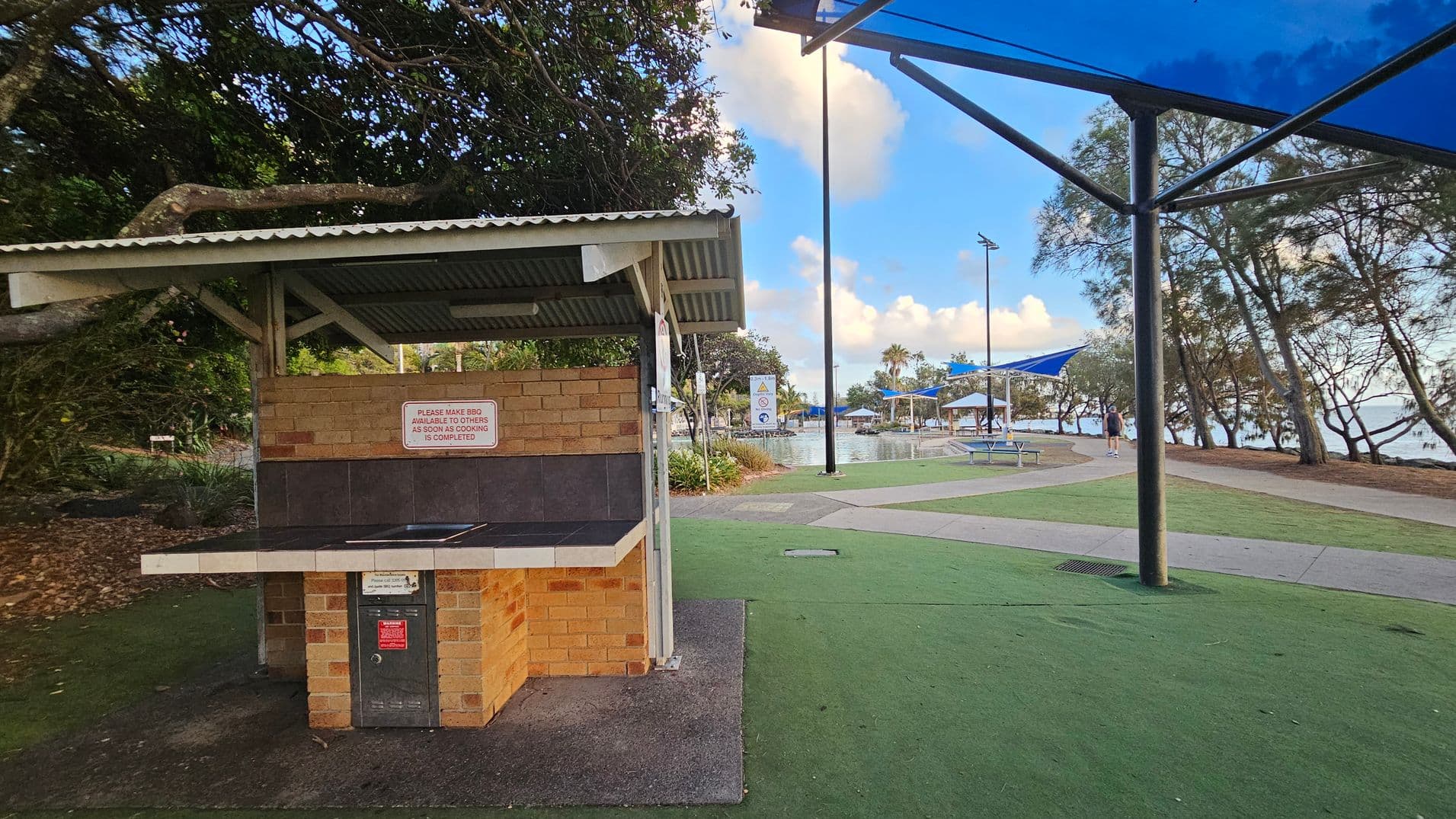 A public barbecue area with a brick structure and a metal roof on the left. A sign on the barbecue instructs users to make it available to others after use. The ground is covered with artificial grass. On the right, there are pathways, picnic tables with blue shade structures, and trees. In the background, a body of water is visible, along with more trees and a clear sky.