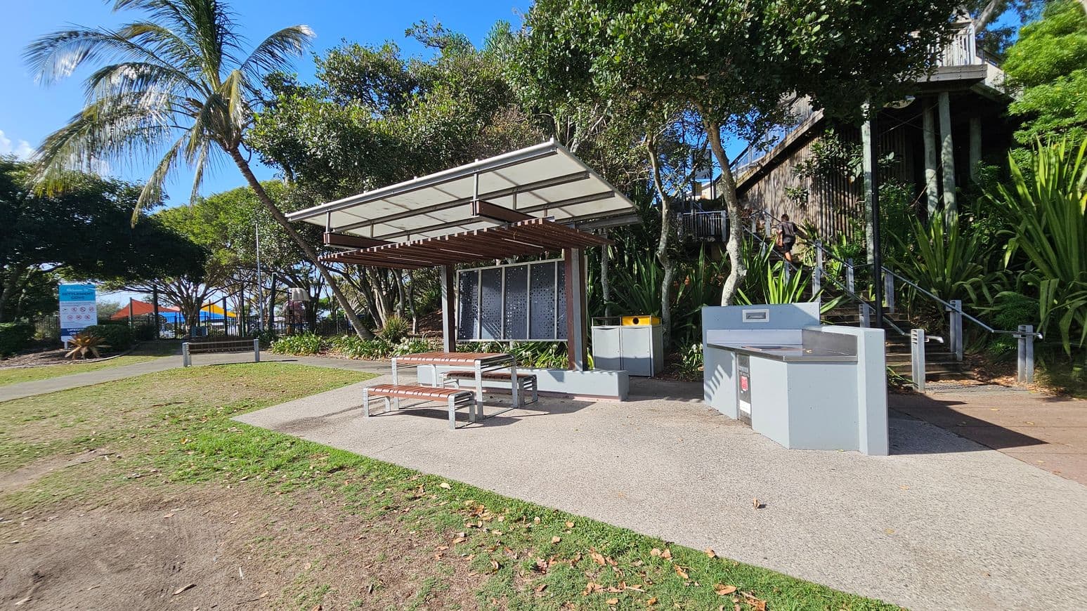 A park setting with a covered picnic area in the centre, featuring a metal and wood structure with benches and a table. To the right, there is a modern barbecue station. In the background, lush green trees and plants surround the area, with a staircase leading up to a higher level. On the left, a sign and colourful shade structures are visible, possibly indicating a playground. The ground is a mix of grass and paved paths.