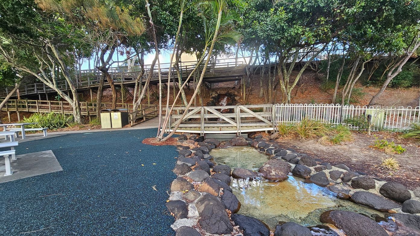 A park area with a small pond in the foreground, bordered by large dark rocks. To the right, there's a white metal fence and some low plants. In the centre, a wooden bridge crosses over a small stream. Behind the bridge, a wooden walkway is elevated among tall trees. The ground is covered with a mix of blue rubberised surface and concrete, with picnic tables on the left. The area is shaded by dense tree foliage.