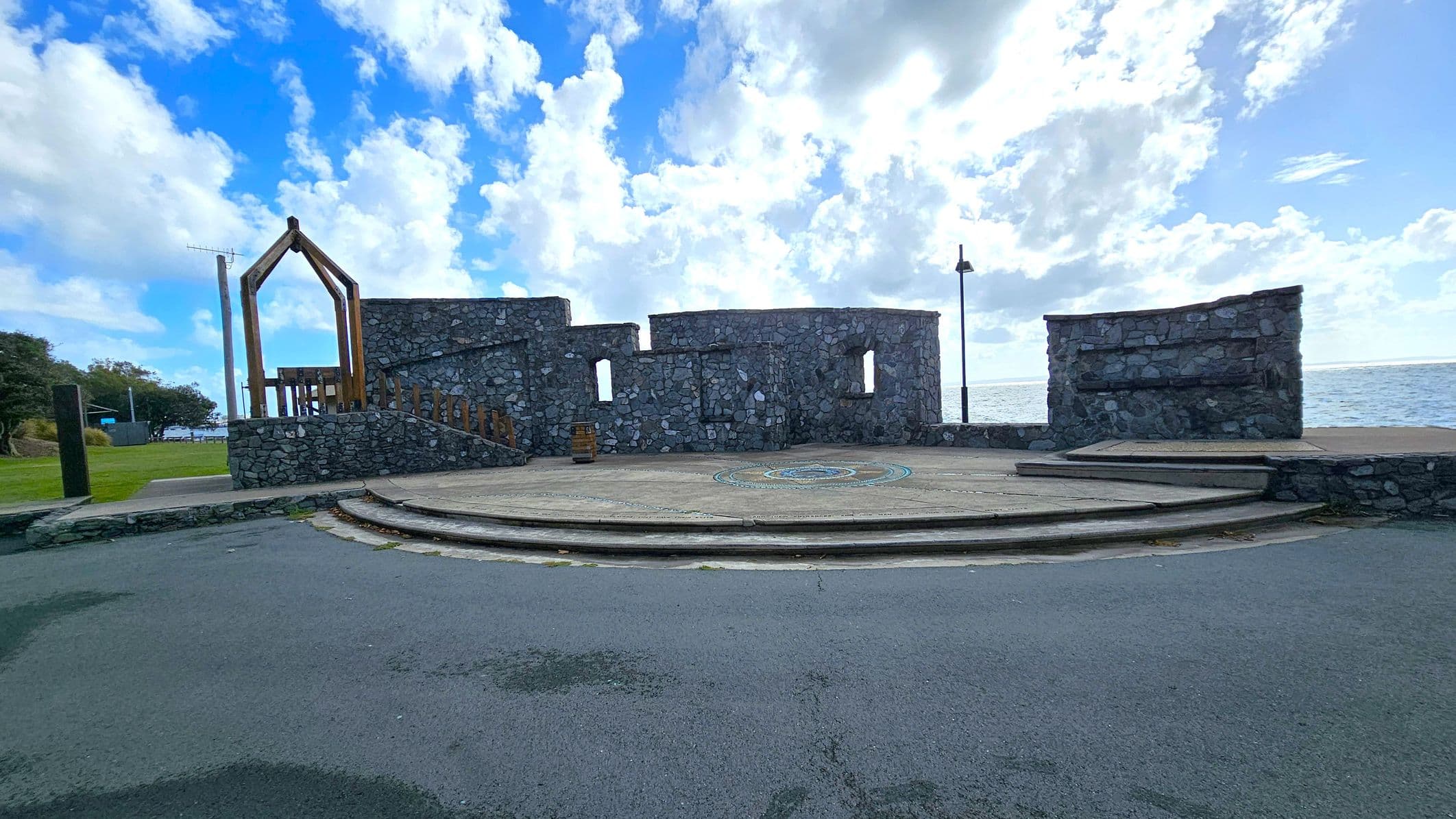 A stone amphitheatre with a circular stage in the centre, featuring a colourful design. The structure has several stone walls with rectangular openings. On the left, there is a wooden frame structure and a grassy area with trees. The right side shows the sea in the background. The foreground has a paved area leading up to the stage.