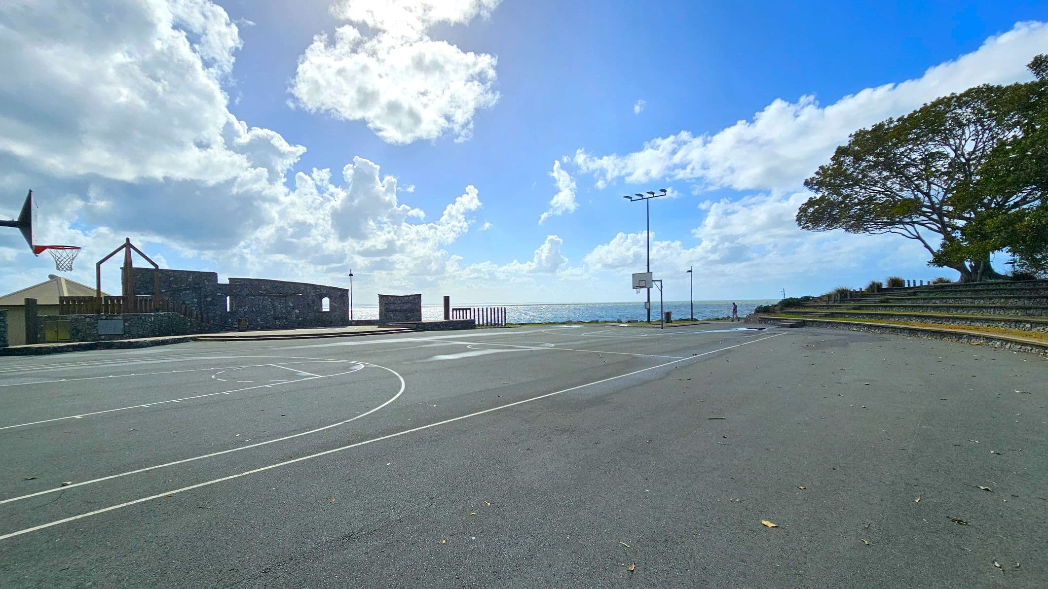 An outdoor basketball court with two hoops, one on the left and one in the centre. The court has white lines and a smooth surface. On the left, there is a stone structure with a wooden element and a small building. In the background, the sea is visible under a partly cloudy sky. On the right, a large tree provides shade over tiered seating. A person is walking near the sea in the distance. The ground is scattered with a few leaves.