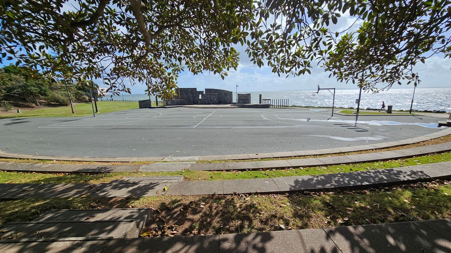 A basketball court is in the centre, with a hoop on the right. The court is surrounded by a grassy area with trees, casting shadows on the ground. In the background, there's a stone structure near the water's edge, and the sea is visible beyond. A person is walking along the waterfront path. The foreground has a paved walkway with grass and fallen leaves.