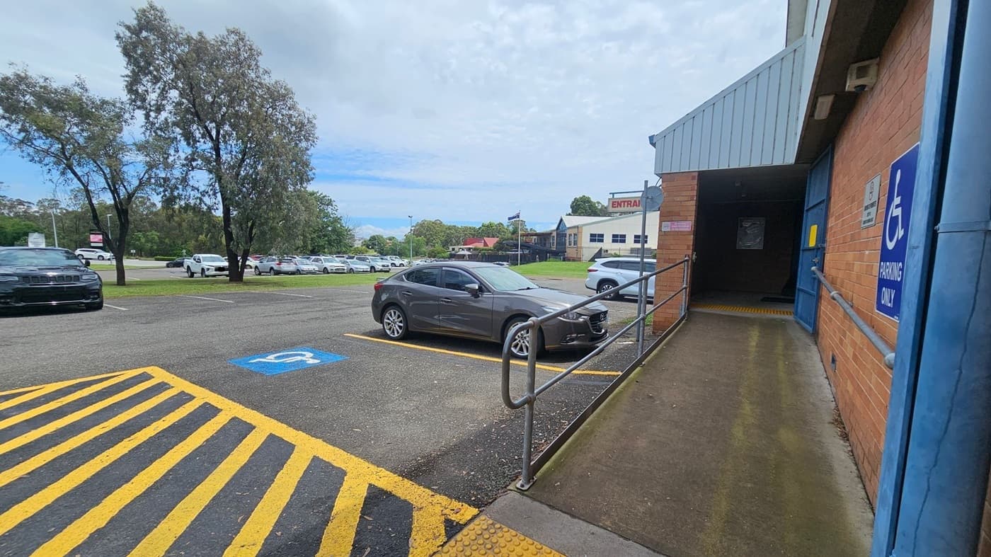A car park with a grey car in a designated disabled parking space marked with a blue sign. Yellow-striped lines are painted on the ground. On the right, a brick building with a ramp leading to a door, also marked with a disabled parking sign. In the background, several trees and more parked cars are visible, along with a building with a sign that reads "ENTRANCE".