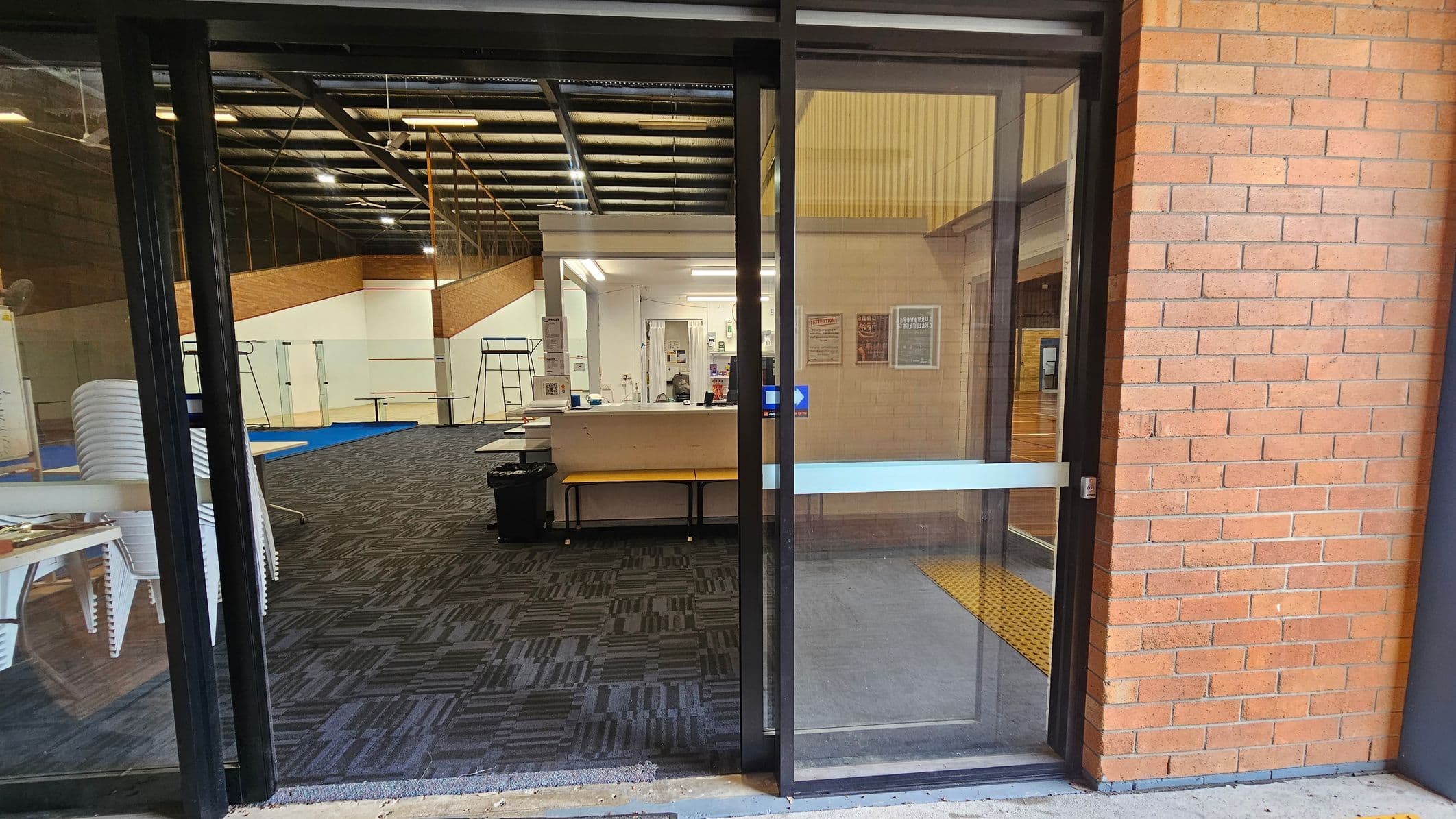 A view through a glass door into an interior space. On the left, there are stacked white chairs and a blue mat on a patterned carpet. In the centre, a reception desk with a computer and various notices on the wall behind it. The ceiling has exposed beams and lights. On the right, a brick wall with a yellow tactile paving strip on the floor near the door.
