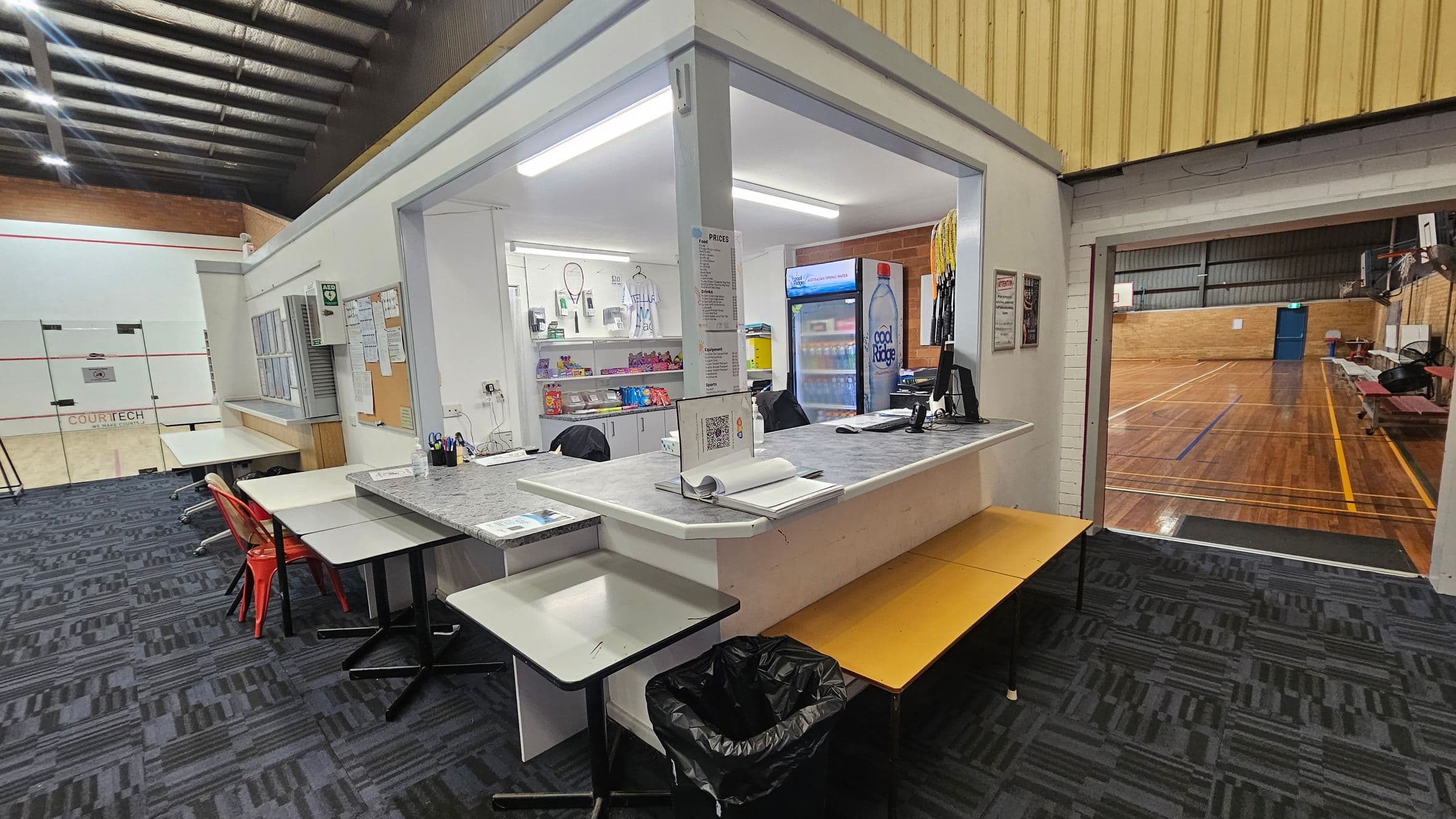 A sports facility interior with a reception area in the centre. The counter has a computer, stationery, and a price list. Behind it, shelves display sports equipment and snacks. To the left, a squash court with glass doors is visible. The floor is carpeted with a dark pattern. On the right, a wooden-floored basketball court with benches and a hoop. The ceiling is high with exposed beams and lighting.