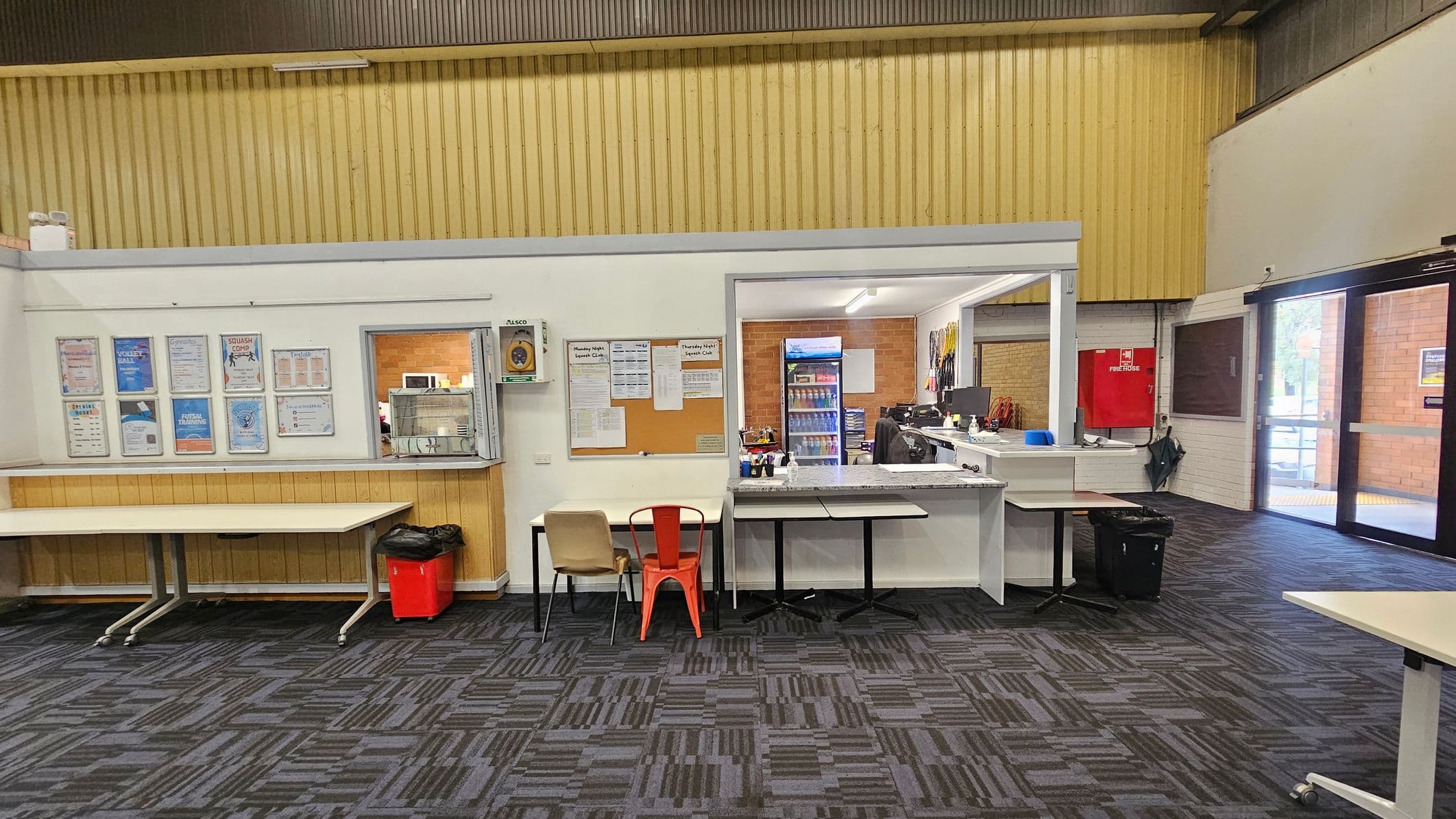 A room with a patterned carpet floor and a high ceiling. On the left, there are tables against a wall with various posters. In the centre, a small service window with a counter and two chairs. To the right, an open area with a desk, computer, and a fridge stocked with drinks. A red fire hose box is mounted on the wall. Large glass doors on the far right lead outside, allowing natural light to enter.