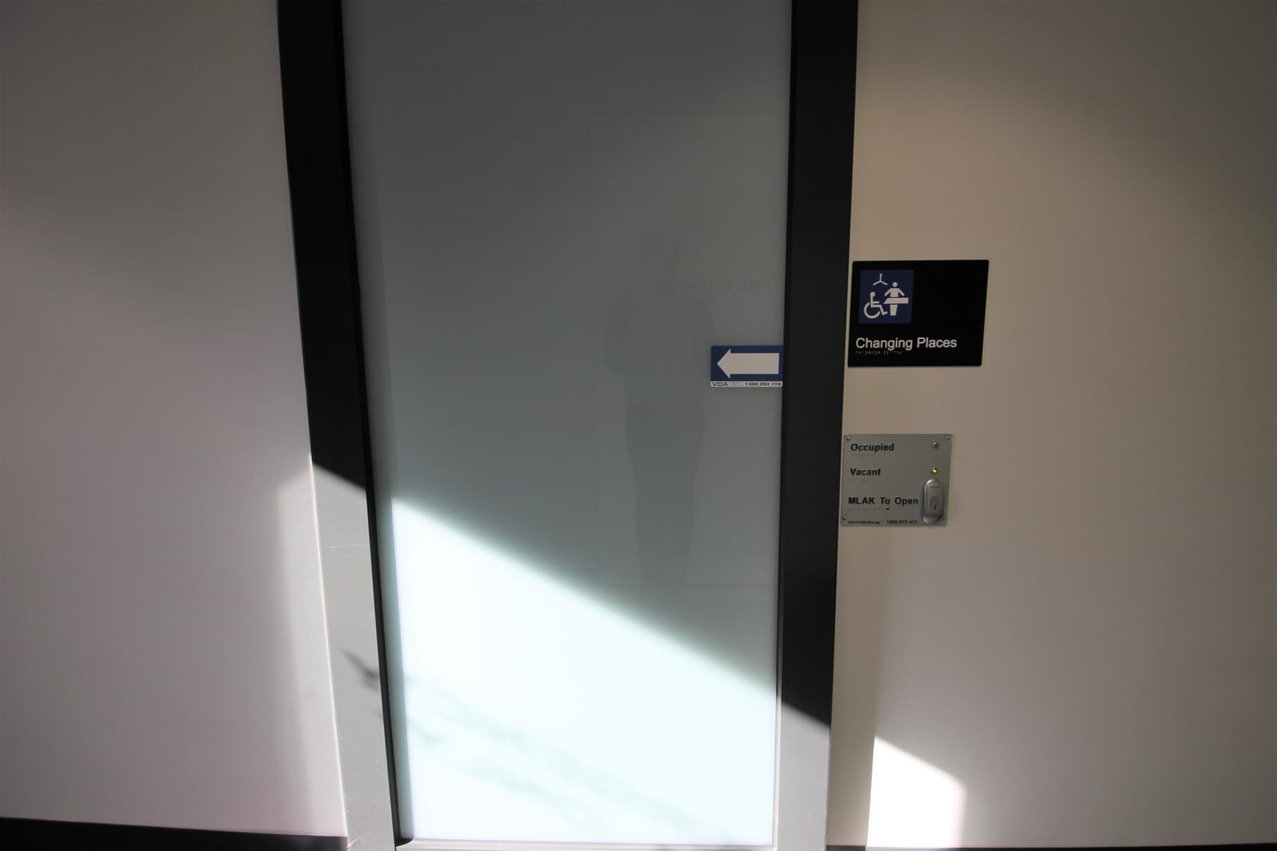 Changing places toilet at Somerville Recreation Centre. Changing places have an electronic sliding door with a push button for opening and different notification lights for occupied and vacant. A white arrow within a blue small rectangular board can be found on the grey door. There are black borders on the sides of this door.