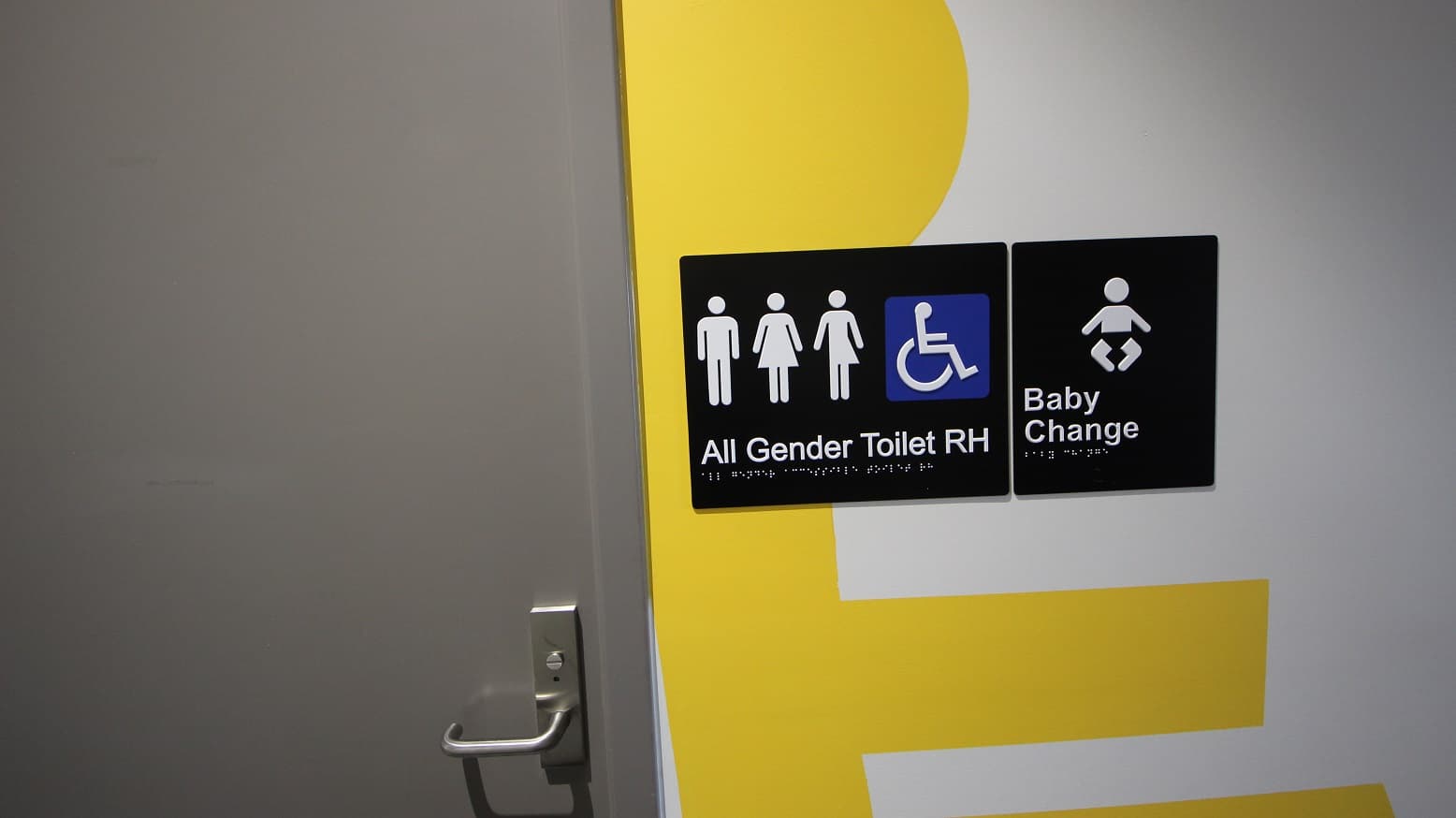 An 'All Gender Toilets' and baby change at Somerville Recreation Centre. The toilet signs have a male, female, non-binary, accessible sign, and baby icon. It has a grey door with a metal handle. There are irregular yellow patterns painted on the white wall.