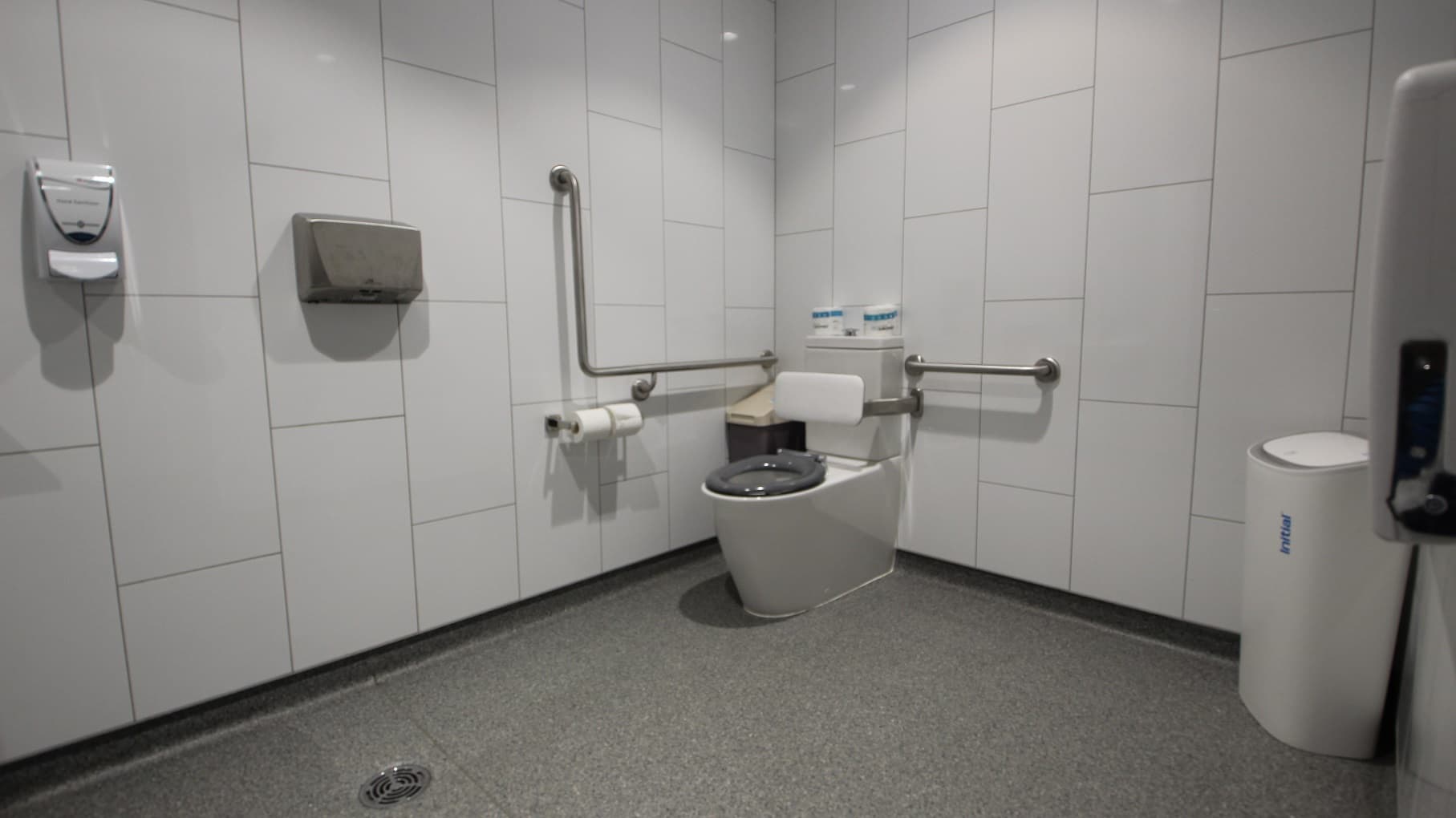 Inside the “All Gender Toilets” is an accessible toilet with a white cushion for back support. On the side of the toilet seat is a brown rubbish bin with a beige lid. There are toilet papers and grab bars mounted on the white tiled wall. There is also a hand dryer and air purifier available.
