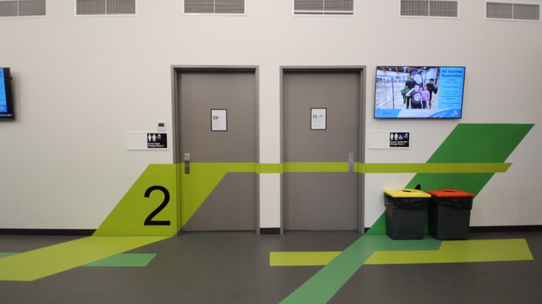 Accessible toilet and change rooms 1 and 2 available on this floor of Somerville Recreation Centre. These two floors have different hues of green paint on some parts of its door and walls. On the right are rubbish bins with orange and yellow lids. A large TV is mounted on the white wall on the right. Several exhaust hoods can be seen on the upper wall.