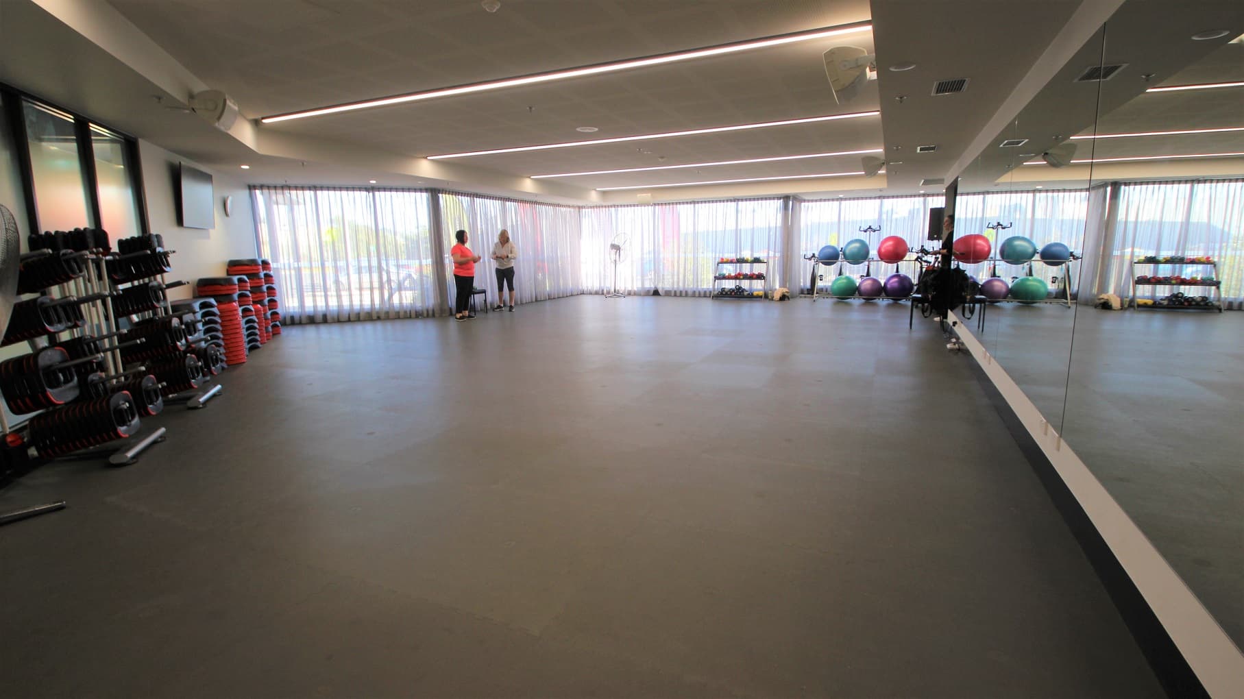 Room for Group Fitness Classes at Somerville Recreation Centre. One of its features is a floor-to-ceiling mirror in front. The glass walls are covered by white thin curtains. Found also in this room a range of weights, exercise balls, and other gym equipment. The floor is covered with a grey mat.