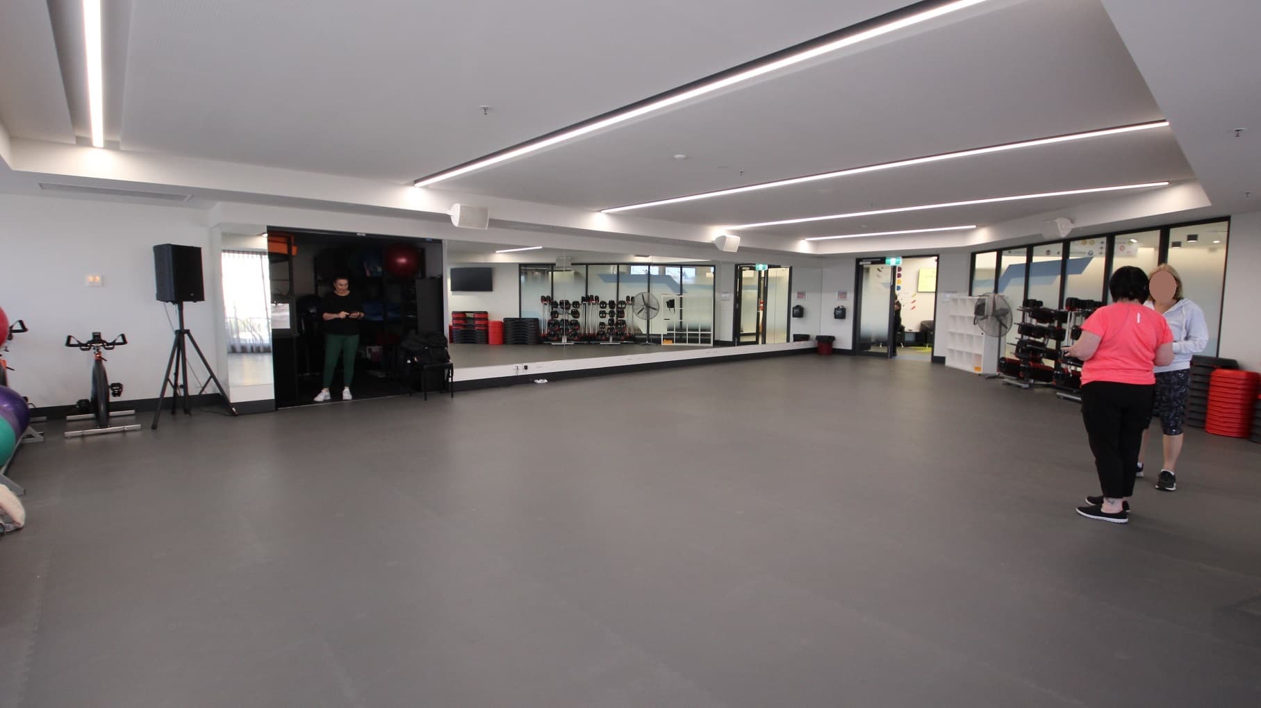 View opposite the entrance door at the Group Fitness Classes room at Somerville Recreation Centre. There is a floor-to-ceiling mirror in front and next to it is a dark room and a white cabinet for bags near the glass doors. Near the gym balls are a bicycle and a speaker with a stand.