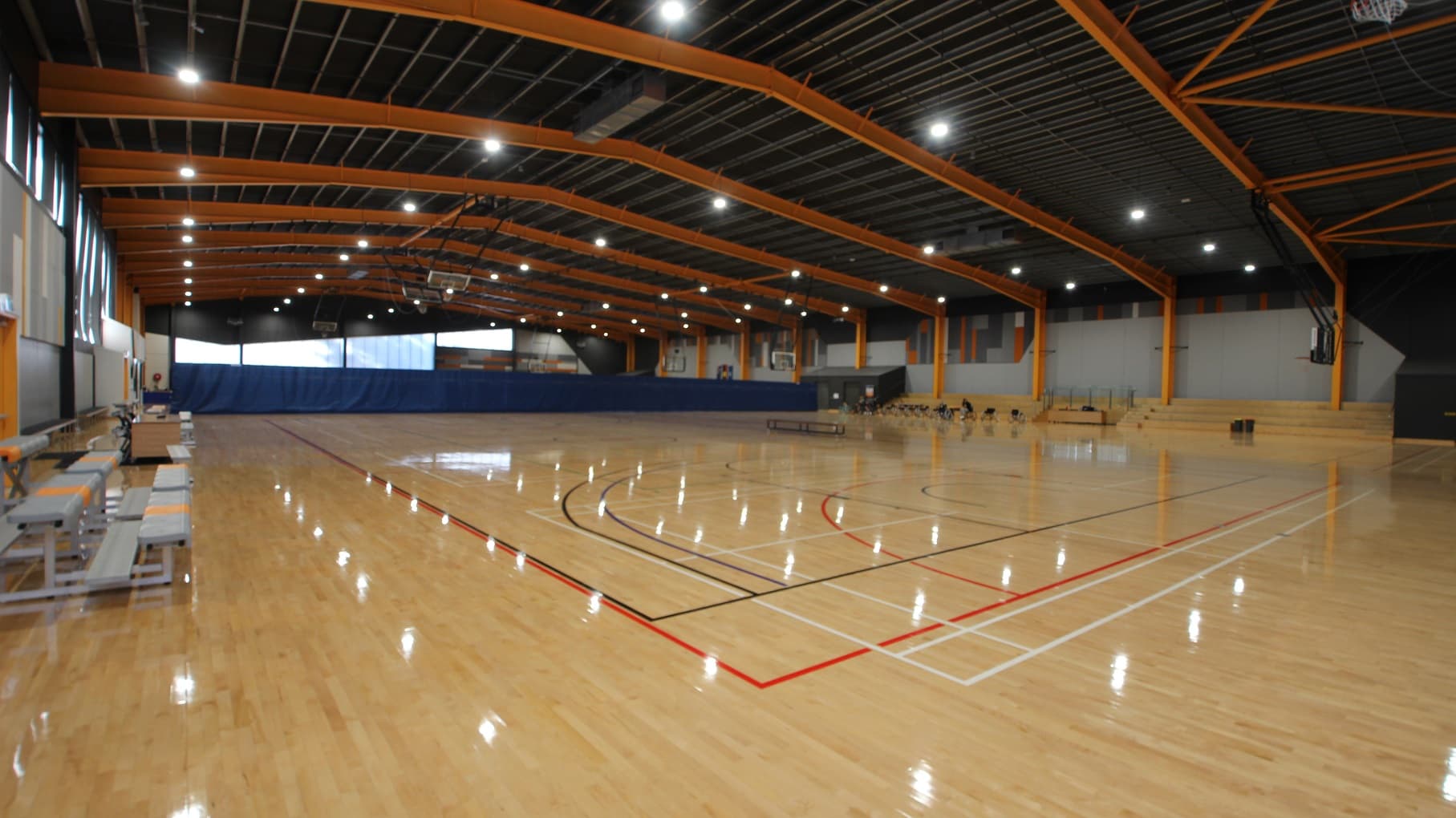 The lights coming from the fluorescents on the ceiling are seen to be reflected on the polished pine timber floor. The scorer's table is located in between the grey benches with orange lanes on the left side of the court, while on the opposite is a 4-tier seating. The stadium has a grey ceiling and bright yellow metal archways.