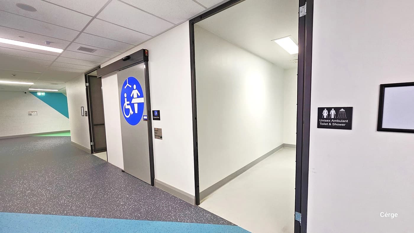 The sign outside the lobby indicates "Unisex Ambulant Toilet and Shower" and has a blank frame beside it. To the left, another sign designates the bathroom is accessible. The floor features grey and blue granite-style tiles, and the wall is painted with white and grey accents. Further to the left is an open door leading to another bathroom.