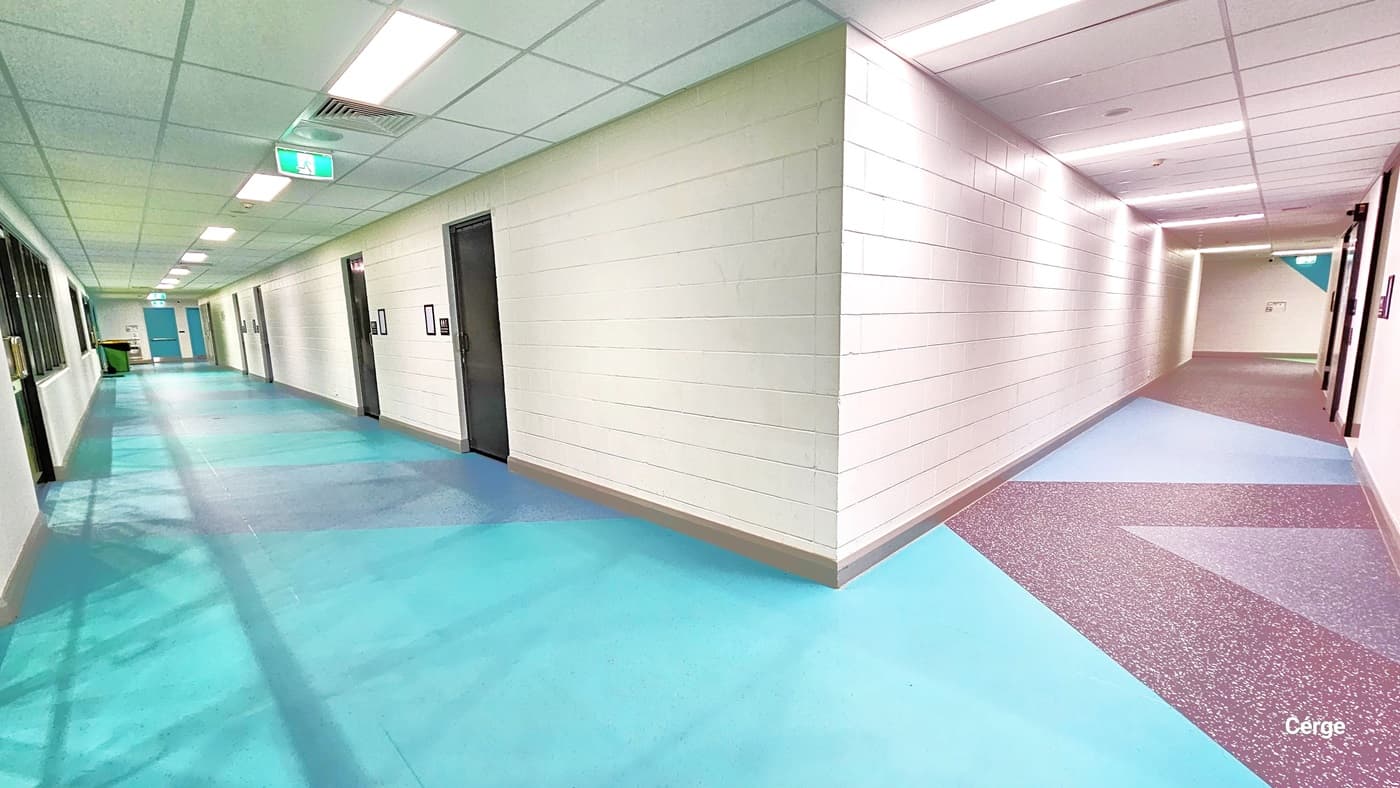Lobby leading to the change rooms. To the left, the floor has a granite design with pink and grey "V" patterns. The white brick design wall complements the bright ceiling lights. To the right, the floor combines green and grey granite designs. The ceiling is illuminated by a row of fluorescent lights. Many brown doors leading to the changing rooms are visible, and further ahead, two green doors with stainless steel handles stand out.