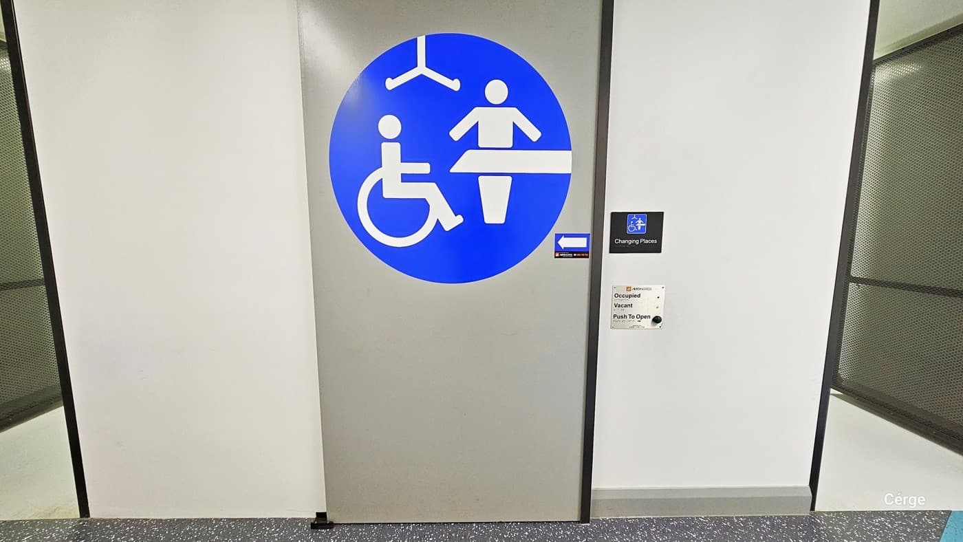 The sign on the wall designates this is an accessible bathroom, with white text in a blue circle. An arrow to the left points to the "Changing room." The walls are white, and the stainless steel doors on both sides feature a mesh-like design.