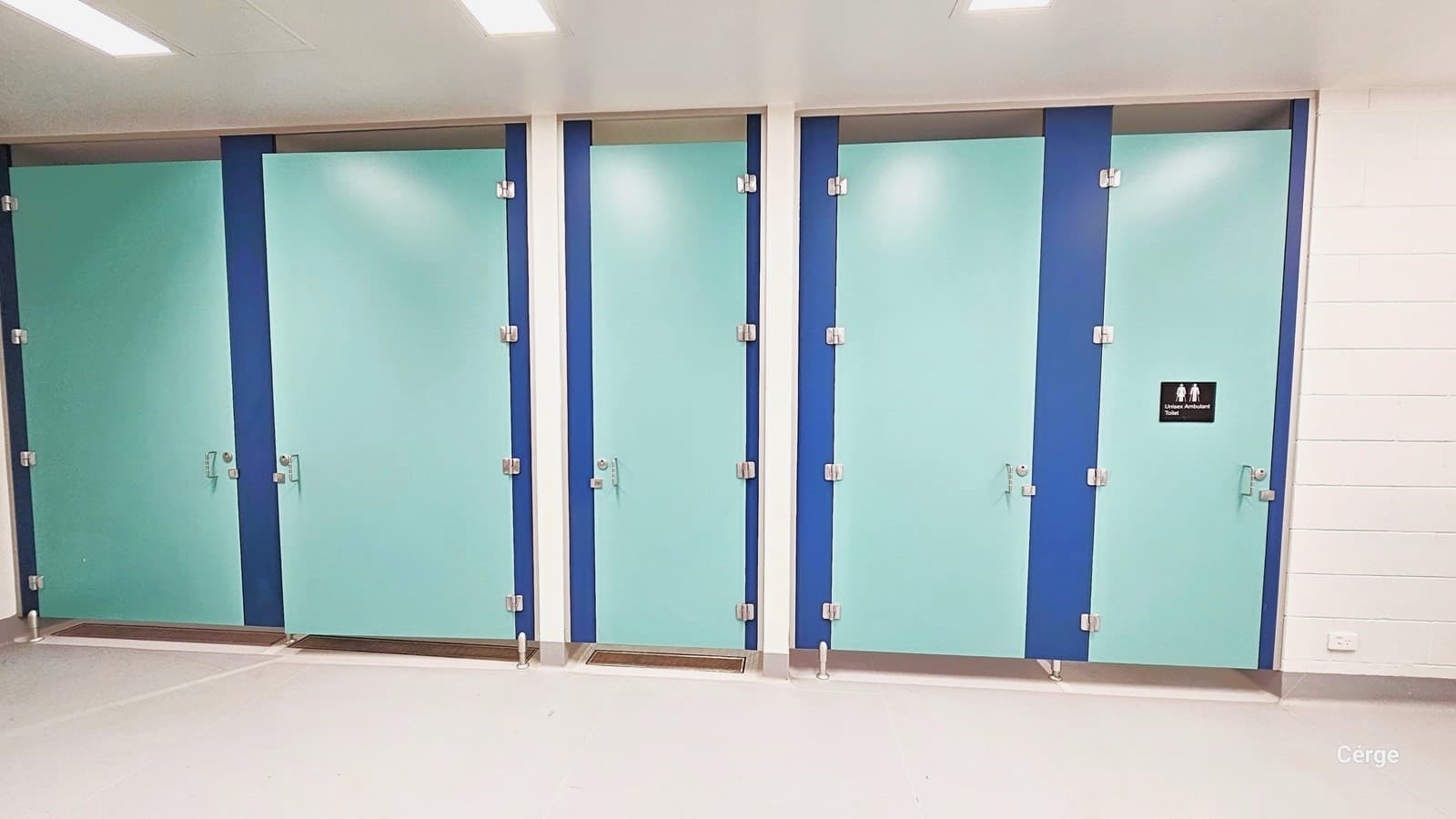 In this area, there are five bathroom stalls with doors painted in light green and blue. The last right door is indicated as ambulatory. The last two left doors have larger dimensions. The room is well-lit with multiple fluorescent lights, featuring white brick-like finished walls and a matte white floor.