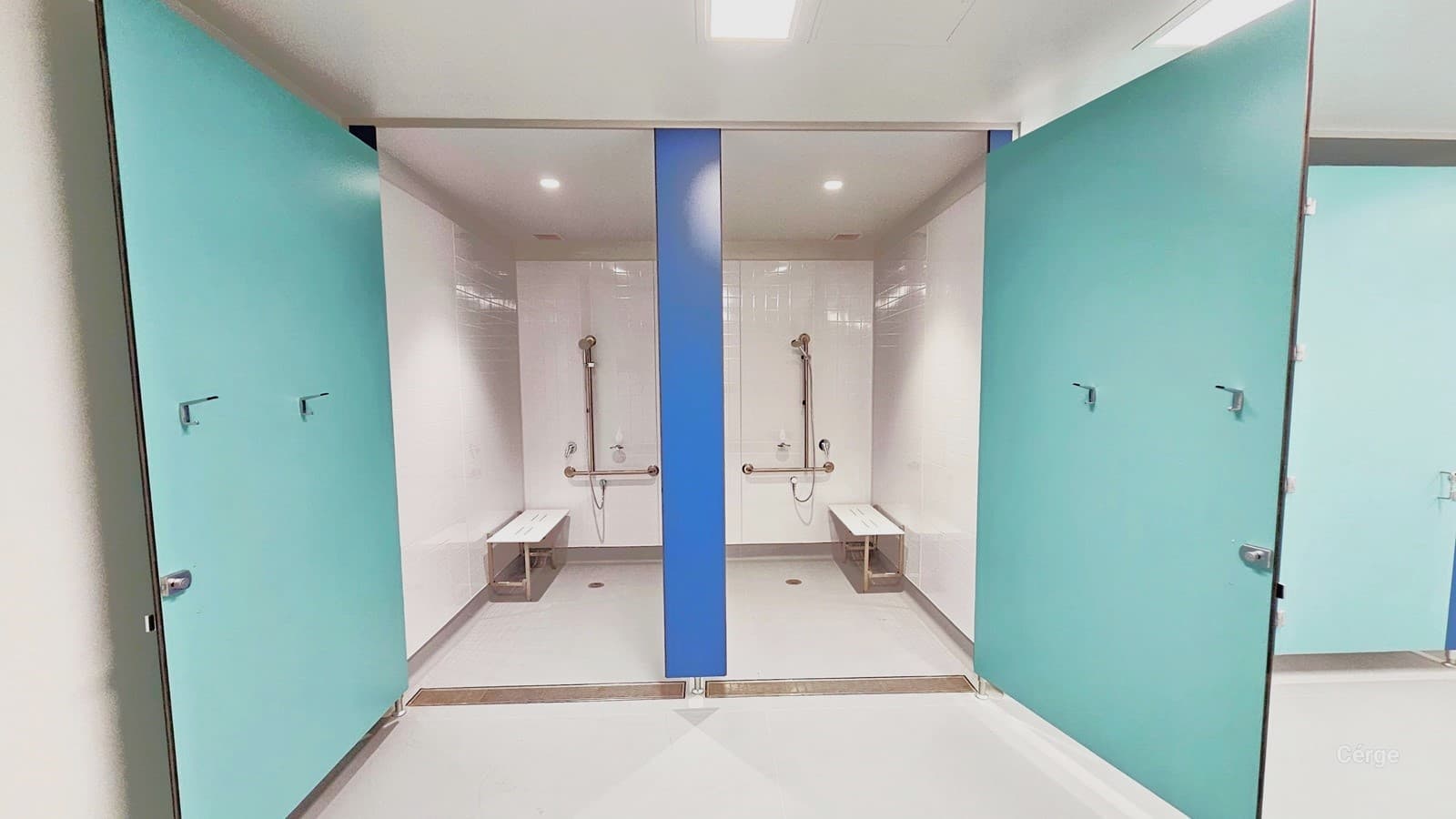 This is the interior of a larger bathroom stall. These two stalls are furnished with stainless steel showers and white wall-mounted handrails. Each stall has white bathroom benches. The walls, floor, and ceiling are all white, while the door is painted in light green.
