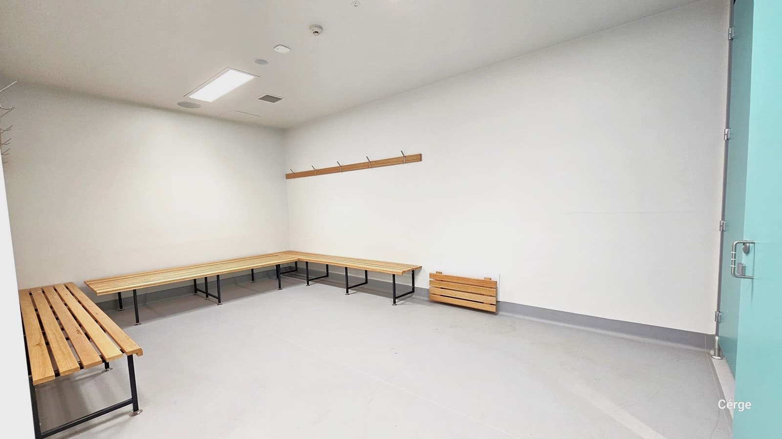 This is the change area. The room is painted entirely in white, including the ceiling and walls. The floor is light grey, and the room is brightly lit by overhead fluorescent bulbs. It features centralized air conditioning on the ceiling. Wooden benches and a wooden panel for hanging items are present.