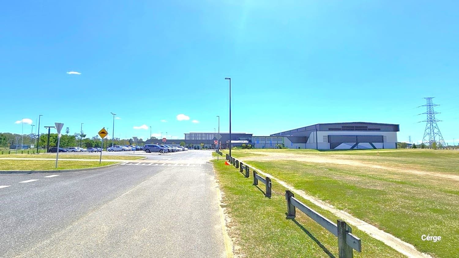A wide road leads towards a large, modern building in the centre, with a car park to the left filled with vehicles. On the right, there's a grassy field bordered by a wooden fence. Several street lamps and traffic signs are visible along the road. In the background, a tall electricity pylon stands to the right, and trees line the horizon on the left. The sky is clear and blue.