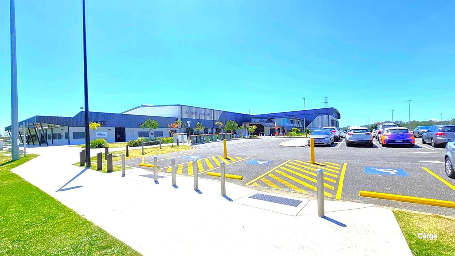 A modern building with a dark roof and light walls is in the centre, surrounded by a well-maintained garden with small trees and shrubs. To the right, a car park is filled with various vehicles, including a blue and a white car. Yellow lines and disabled parking symbols are visible on the asphalt. On the left, a wide concrete path runs alongside a grassy area. Tall lamp posts are positioned along the path. The sky is clear and blue.