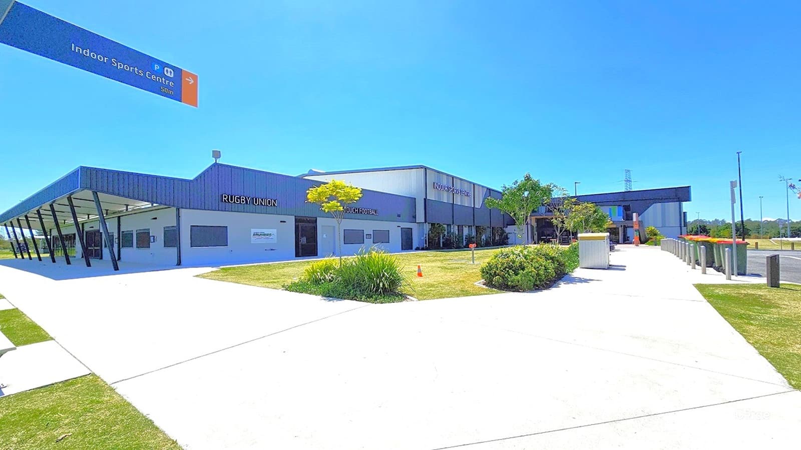 A modern sports facility with a sign for "Rugby Union" on the left side of the building. The structure has a grey and white exterior with a covered walkway supported by black pillars. In the foreground, there is a wide concrete path bordered by grass and small shrubs. A sign overhead points towards the "Indoor Sports Centre" 50 metres away. The area is well-maintained with a few small trees and bushes along the path.