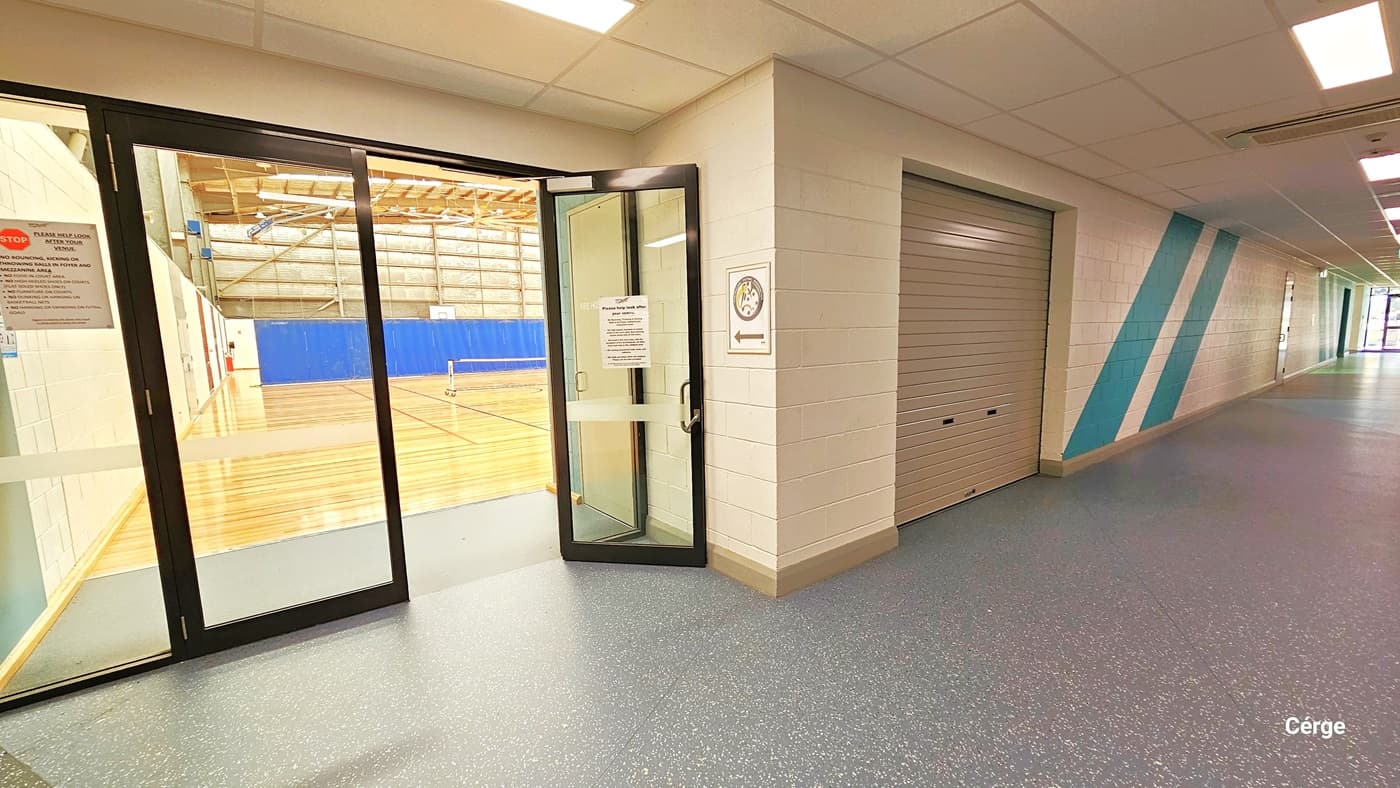The area is well-illuminated by fluorescent lights on the cream ceiling. To the right, the lobby has grey granite design flooring and a brick design wall with green diagonal accents. An open glass door leads to the court, and there's another entrance to the court through a stainless roll-up door with a darker cream shade.