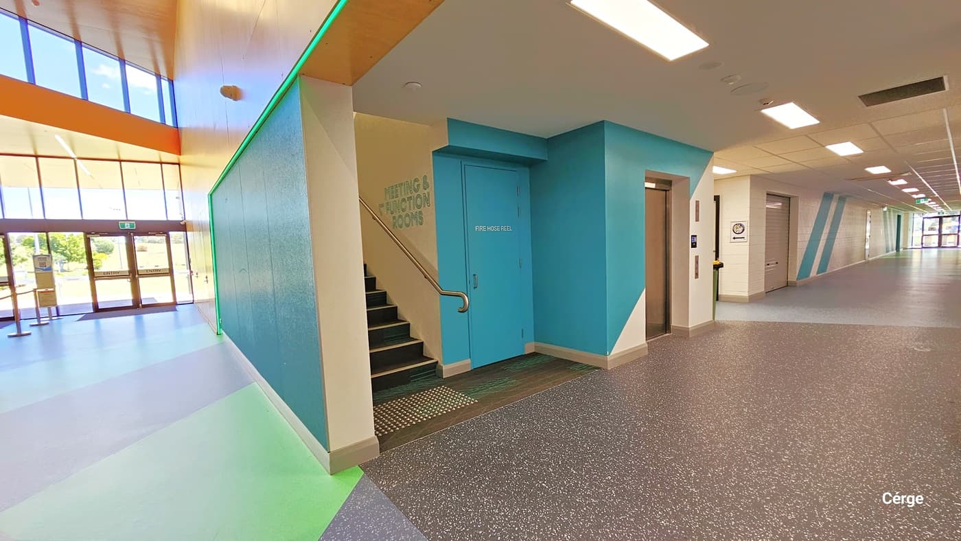 This is a spacious corridor with glass windows at the end. To the left, stairs lead to Level One, accompanied by a lift. Further left, there's access to Courts Four and Five. The area is brightly lit by fluorescent lights on the ceiling. The left side features multi-colored flooring, while the right side has grey granite design flooring.
