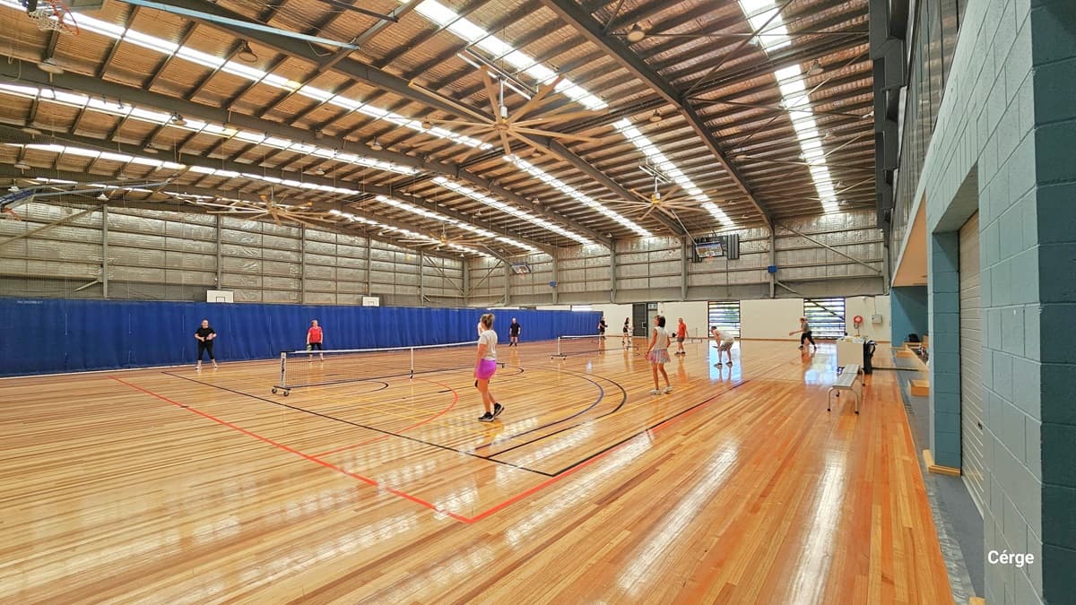 Entering Court 4. The area is brightly lit with long strips of fluorescent bulbs on the ceiling. The brown ceiling has a corrugated finish and is adorned with a couple of large brown fans. The timber floor is marked for various ball games. At the far end, there are glass windows and a large flatscreen on the white brick design wall. Several people are engaged in ball games, and there's a white table with bench chairs in the corner.