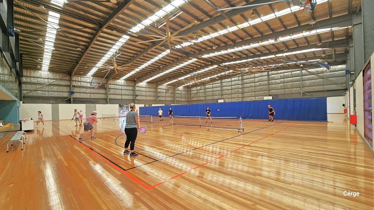 Another view of the court. The area is well lit by the long strips fluorescent bulb from the ceiling. The ceiling is painted brown and has corrugated finish, accented by a couple of large brown fans. The timber floor has markings intended for different types of ball games. The far end shows a blue cloth divider. There are numerous people playing badminton. A white table and white bench chairs are also positioned on the left.